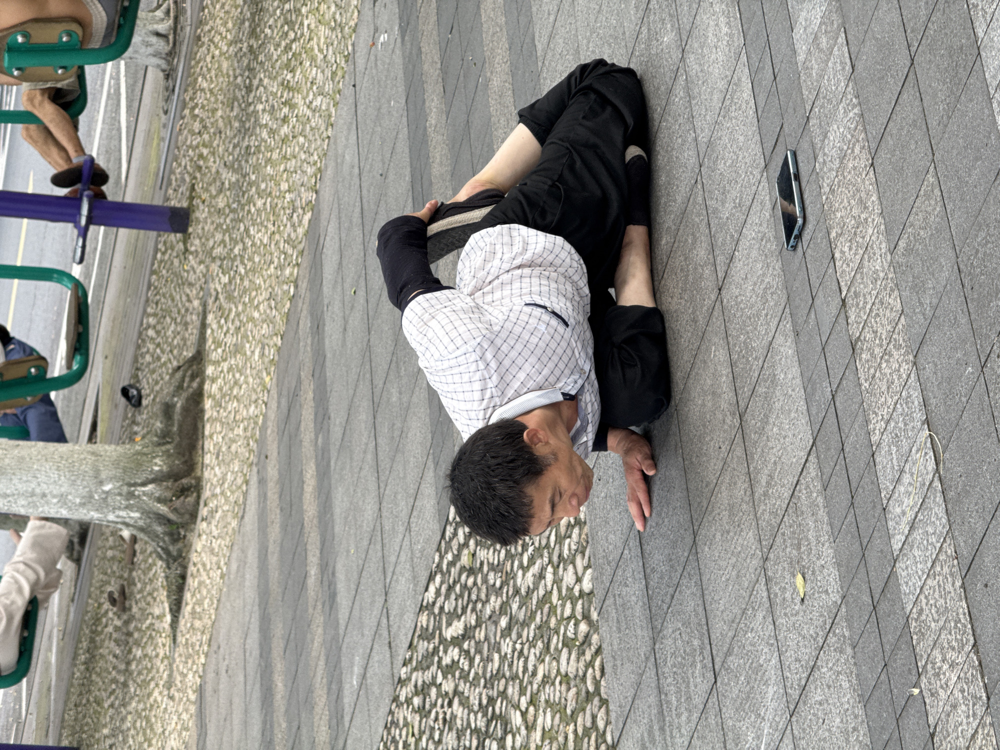 An East Asian man is depicted performing a stretching or yoga pose on grey tiled pavement in the city of Jiangjin, China. He is positioned on his knees, with his torso lowered and his hands flat on the ground in front of him. His head is downcast, and his mouth is slightly open. His legs are bent backward, with his feet raised towards his back, suggesting a deep backbend or quad stretch. He wears a white and black checkered short-sleeved shirt, black trousers, and a black sleeve on his left forearm. His hair is short and dark. A black smartphone lies face-up on the pavement to his right, reflecting ambient light. A small yellow leaf is also visible on the pavement nearby. In the background, beyond a section of textured ground covered with light-colored pebbles, a public exercise area is visible. A tree with exposed roots stands to the left, and green and purple metal outdoor exercise equipment is present. Two other individuals are partially visible using this equipment: one with light-colored trousers on the left, and another with bare legs and black shoes on the right. A paved road with a white line is discernible in the far background.