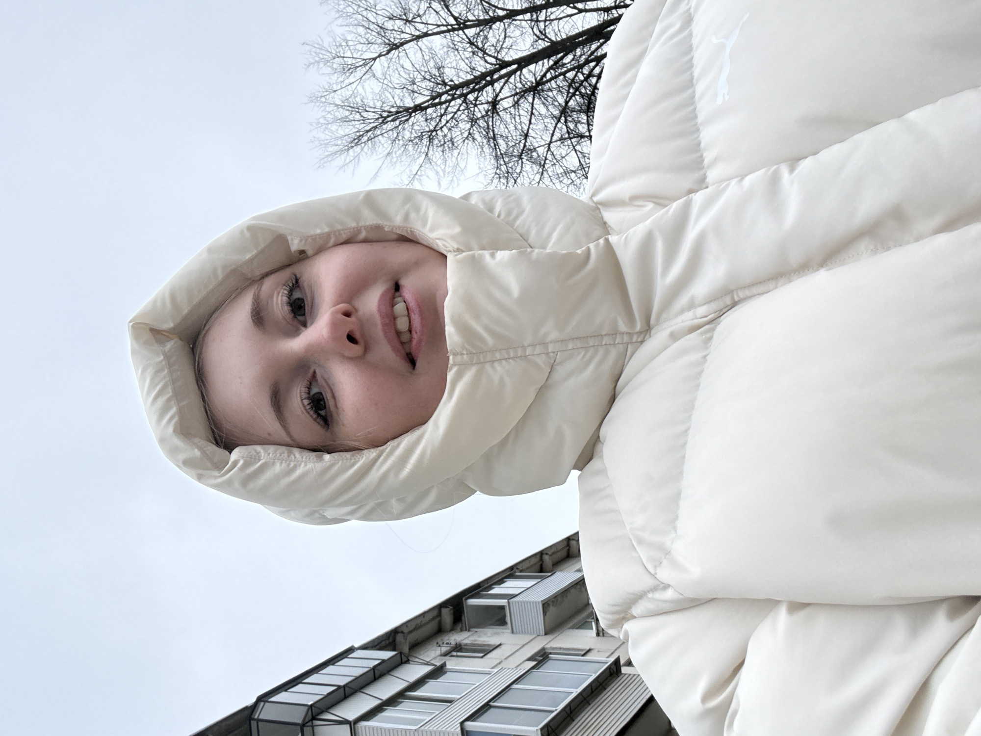 The image is taken from a low-angle perspective, showing a young woman wearing a white puffer jacket with the hood up. She is smiling slightly. In the background, there is a leafless tree against a gray sky. Below the tree and woman, there is a modern building with large, rectangular windows. The image appears to be taken in an urban environment, likely in the city of Unknown, Unknown.