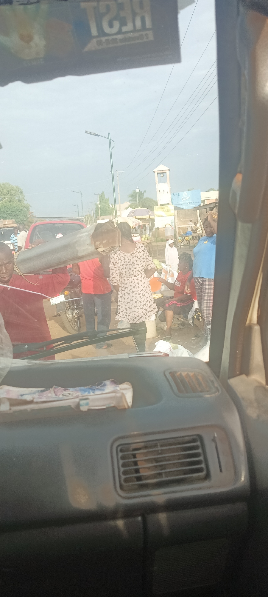 The image, taken from inside a vehicle, captures a street scene in Mangu Town, Nigeria. In the foreground, the vehicle's dashboard is visible. Through the windshield, a group of people can be seen engaged in various activities. A man in a red shirt and another individual are carrying a long, cylindrical object over their shoulders. A woman in a patterned dress stands nearby. Other people are sitting or standing on the side of the road. A red car and a motorcycle are parked in the background. Street vendors are visible under umbrellas or makeshift stalls. A clock tower is visible in the distance.