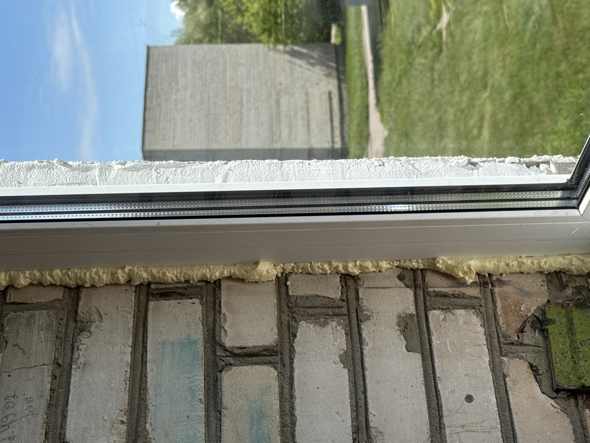 The image captures a close-up view of a window frame being installed against a brick wall. The left side of the frame is bordered by rough, light-colored bricks, some of which appear to have faint blue markings and possibly some graffiti. Expanding foam insulation is visible between the brickwork and the window frame, filling the gap. The window frame itself is white, with a visible inner metal reinforcement.
Through the window, a grassy area and a concrete path are visible. In the background, a large, light-colored rectangular building with a rough texture stands under a clear blue sky with a few scattered clouds. Trees can be seen behind this building, suggesting an outdoor environment. The scene appears to be in a residential or industrial area, possibly under construction or renovation. The overall impression is one of an urban or semi-urban setting on a bright, sunny day. Given the context provided for "Яромінскі сельскі Савет, Belarus", this scene likely depicts a window installation at a building within this administrative region.