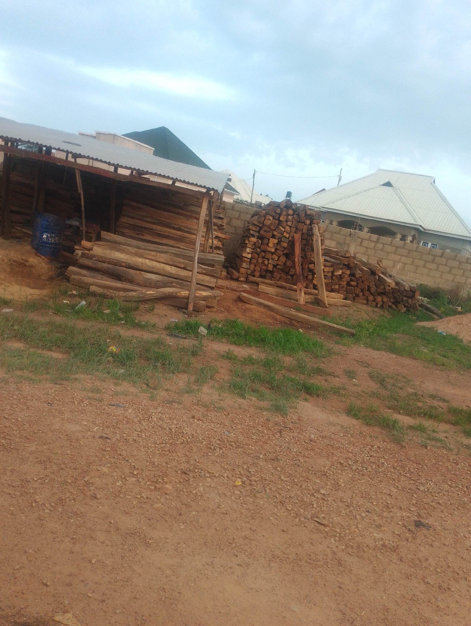 The image shows an outdoor scene with stacks of lumber and a blue barrel, likely at a construction or lumber yard in Kwang, Nigeria. In the foreground, a dirt path slopes upward towards the lumber. Several piles of cut wood are visible, some stacked horizontally beneath a corrugated metal roof supported by wooden posts, and other piles of shorter, more uniformly cut pieces are stacked vertically against a brick wall.
Behind the lumber, a building with a white corrugated metal roof and light-colored walls is partially visible. To the left of this building, a green-roofed structure, possibly another building or a covered area, is also present. The sky is overcast with clouds. The overall impression is of a working area where construction materials are being stored and prepared. There are no people or animals visible in the image.