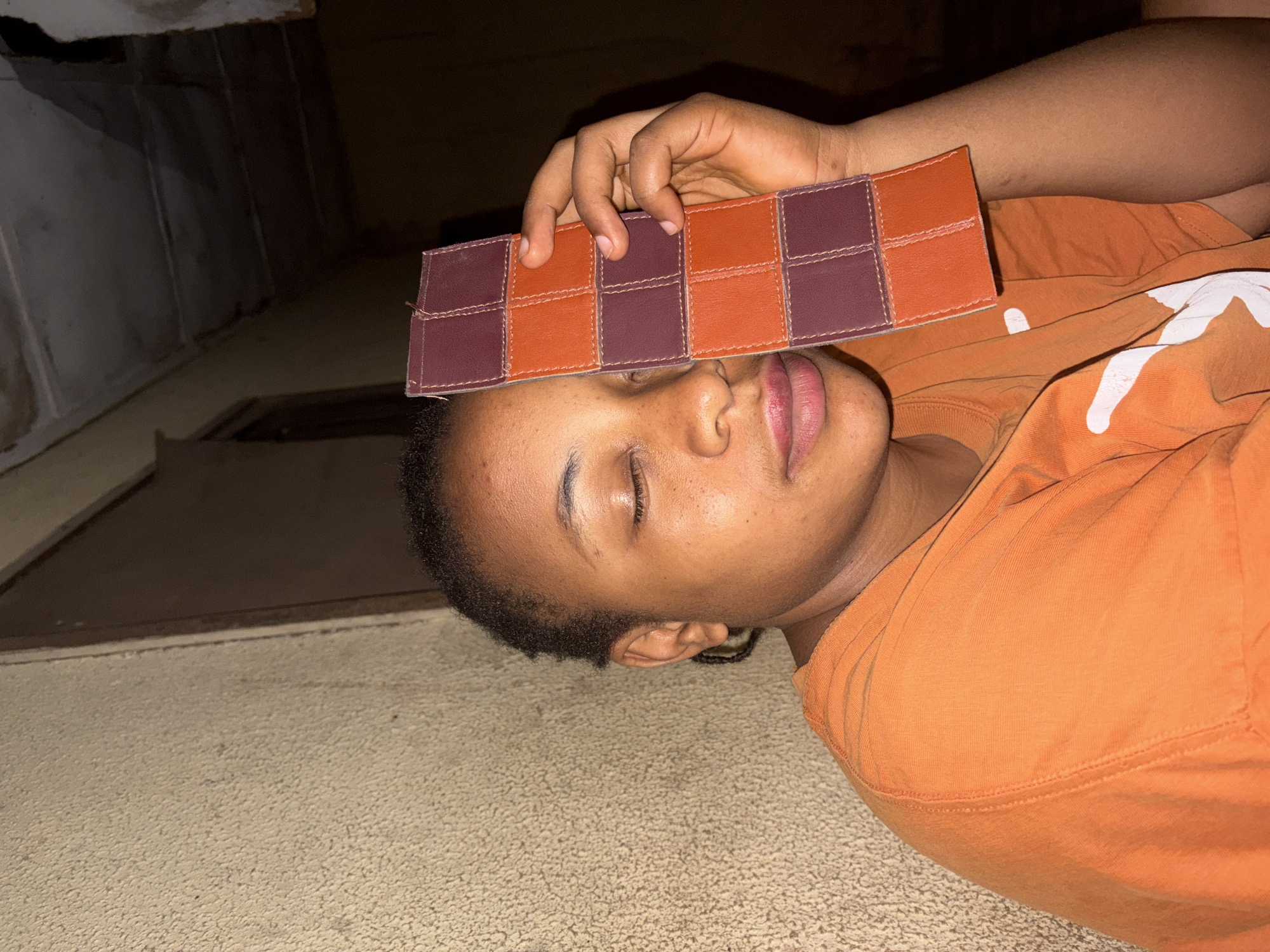 The image shows a person, likely a young adult or teenager, with dark skin and short dark hair, lying on their back with their eyes closed. They appear to be resting or relaxing, with a calm expression and slightly parted lips. The person is wearing a bright orange t-shirt.
One hand is positioned across their forehead, holding a distinctive rectangular object. This object features a checkerboard pattern, comprising alternating squares of dark reddish-brown and orange-brown material, possibly leather or a similar fabric, joined by visible stitching.
The setting appears to be indoors. A light-colored, textured surface, resembling carpet or concrete, covers the floor where the person is lying. The background is darker and less defined, with hints of a wall or ceiling structure. The overall lighting is dim, suggesting an indoor environment, possibly at night or under artificial light. The scene is set in Jos, Nigeria. No visible text is present in the image.