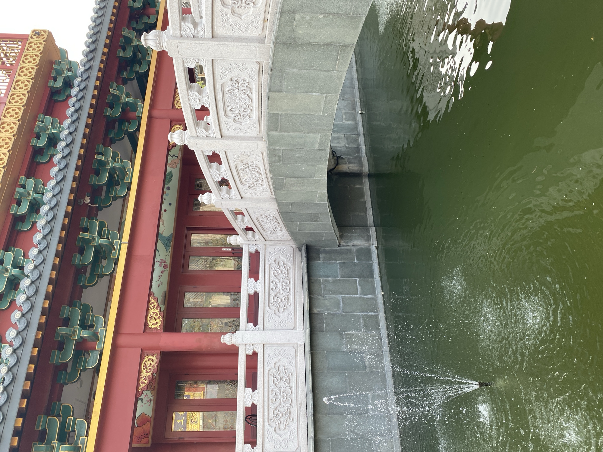 The image depicts a traditional Chinese-style building with red walls and ornate green and gold decorations under the eaves, set against a bright sky. A stone bridge with intricate carvings on its white stone balustrades arches over a body of dark green water. A small fountain sprays water in the foreground, creating ripples on the surface. The water reflects the surrounding architecture. The scene is located at the Tzu Chi Centre in Pantai Indah Kapuk, North Jakarta, Indonesia, a complex known for its Buddhist architecture and gardens. There are no people visible in the image.