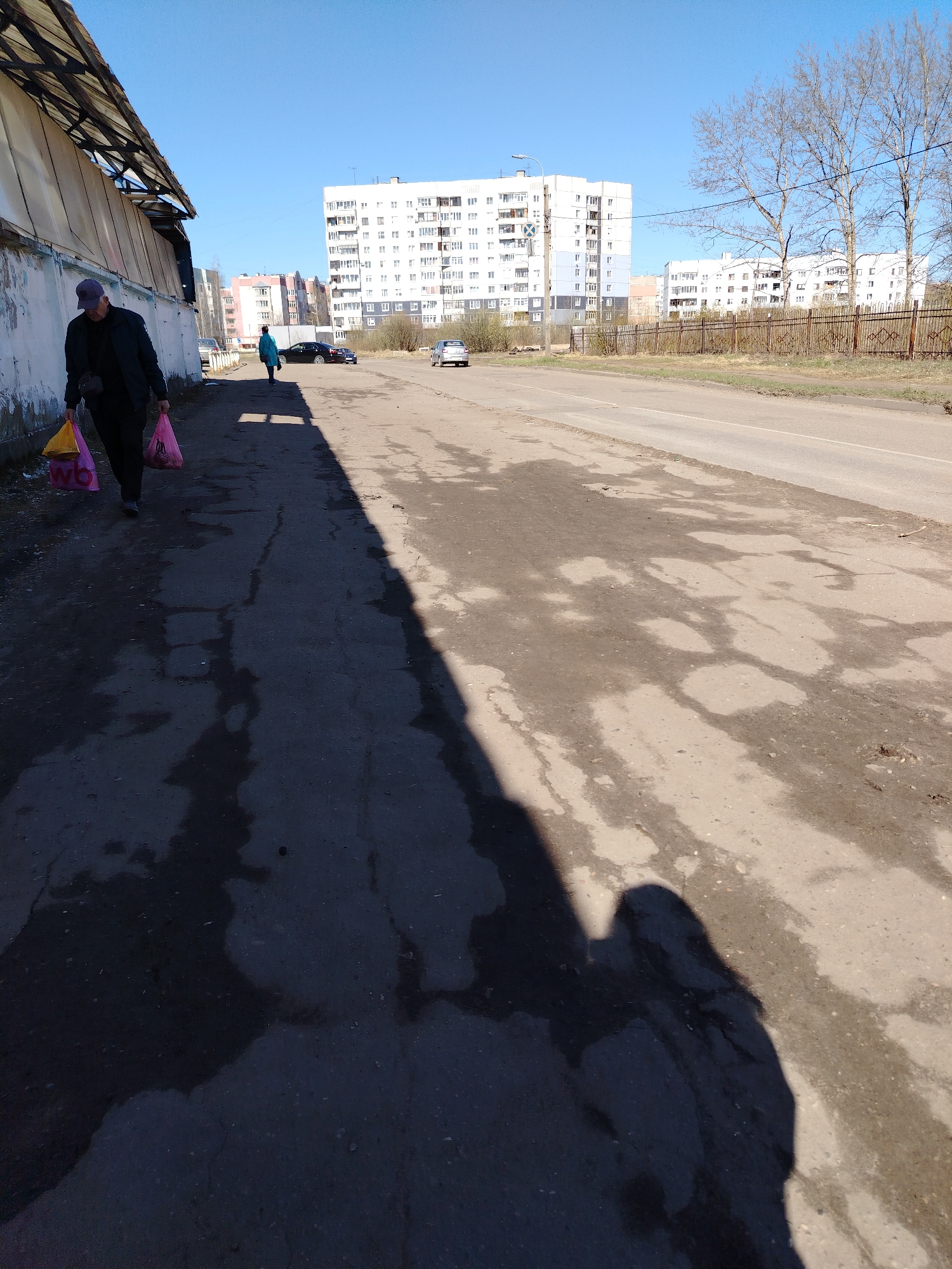 The image depicts a street scene in Rusanovo, Russia, on a sunny day. A person wearing a dark jacket and a blue baseball cap is walking along the left side of the street, carrying two shopping bags, one pink and one yellow with the letters "wb" printed in red. Another person, dressed in a blue coat, is further ahead, walking in the same direction.
Across the street, on the right, is a two-lane road with a rusty metal fence separating it from a grassy area where some bare trees are present. In the background, multiple apartment buildings are visible, mainly white and blue in color.
The pavement is uneven, showing signs of wear and tear, with patches of asphalt and dark, damp spots. A large shadow stretches across the lower part of the image, likely cast by the photographer. The light suggests it is morning or late afternoon. Two cars are parked near the buildings in the distance.
