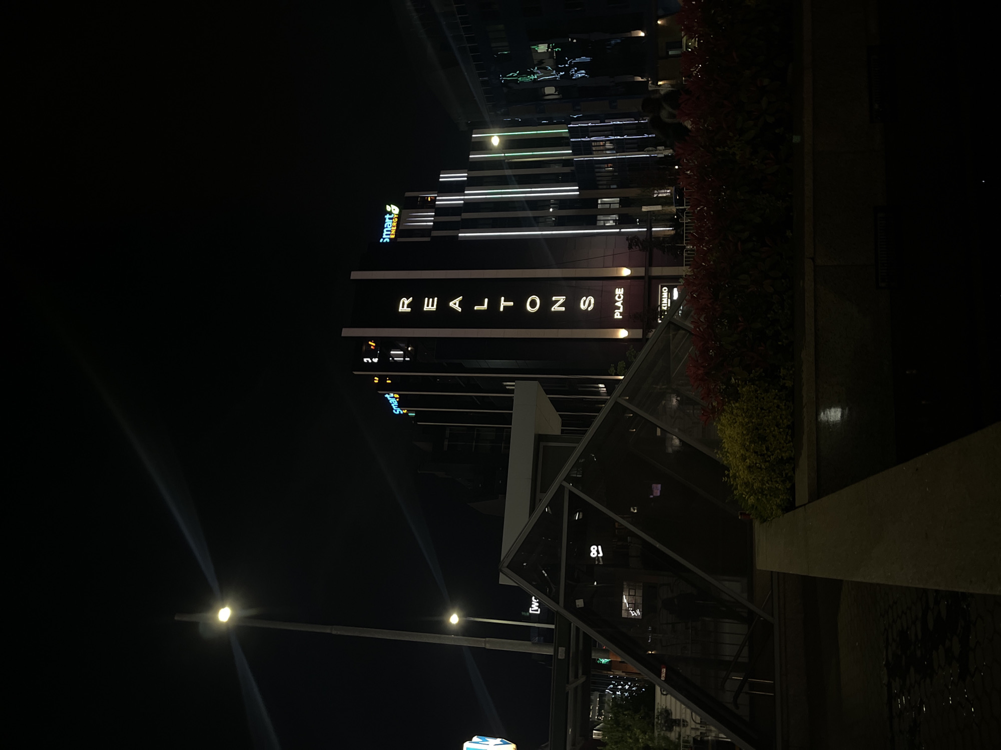 The image presents a modern urban scene at night in Sofia, Bulgaria. The central subject is a tall, contemporary building, dark against the night sky, but accented by numerous illuminated white horizontal and vertical light strips along its facade.
Prominently displayed vertically on the building's side are large, white glowing letters spelling "REALTONS PLACE". Below "PLACE", a smaller, illuminated sign reads "ZIMMO". Further up on the building, a distinct "smart ENERGY" logo, featuring blue and green text, is visible.
In the foreground, to the right, a dense stretch of foliage with reddish leaves borders a dark architectural element or planter. Adjacent to this, there are glass and metal structures, possibly forming part of an entrance or a separate modern building. On the left side of the frame, a bright streetlamp emits a strong, localized light, creating visible rays against the dark backdrop.
The time of day is clearly night, with the sky appearing dark and cloudless. No people are visible, and there is no discernible activity taking place, giving the scene a still and quiet quality despite its urban setting. The overall impression is one of sleek, illuminated modern architecture in a metropolitan environment.