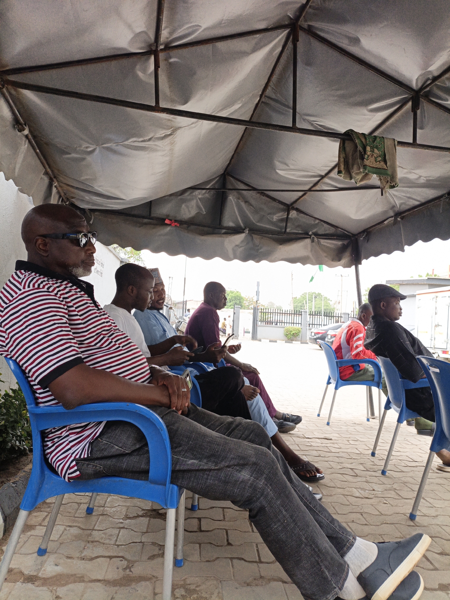 The image depicts a group of six men sitting on blue plastic chairs under a makeshift canopy in Giginyu, Nigeria. The canopy consists of a metal frame covered with a white tarp, from which hangs a patterned cloth. The men are of various ages and are dressed in casual attire, with one man wearing sunglasses and a striped shirt. Some men are looking at their phones. A red car and other men stand in the distance. The area appears to be a waiting place, possibly outside a building. The ground is paved with interlocking stones. A Nigerian flag is visible in the background.