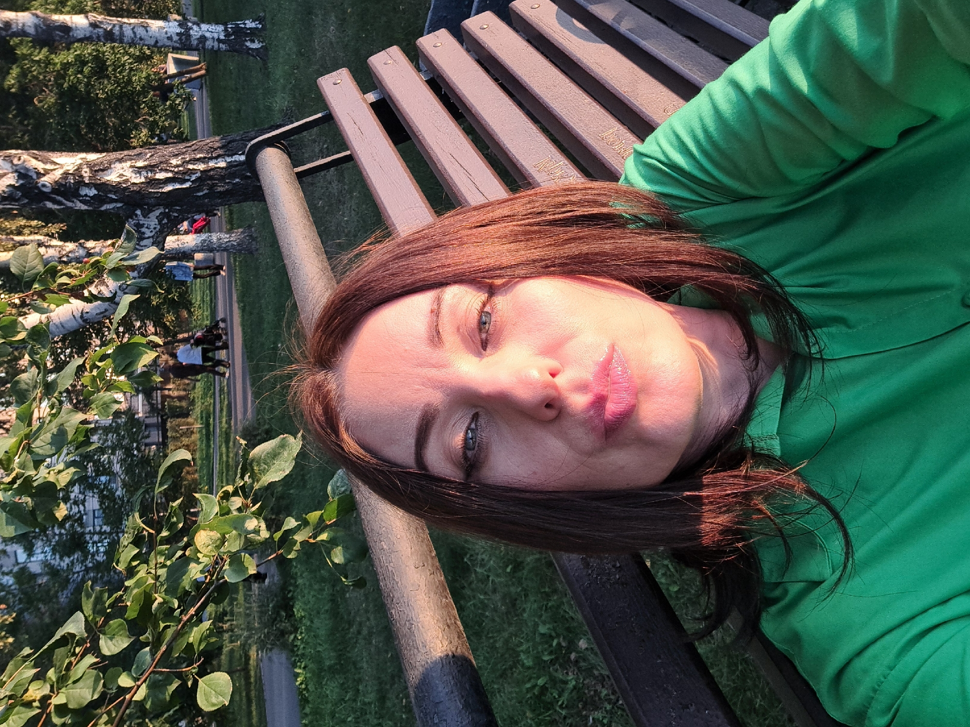A woman with dark brown hair and blue eyes, wearing a green shirt, is taking a selfie while sitting on a park bench. She is puckering her lips. In the background, there are trees, grass, a pathway, and some people walking. The lighting suggests it is either early morning or late afternoon, with warm sunlight casting shadows. The environment appears to be a public park within a city, likely Irkutsk, Russia, given the birch trees common in that region.