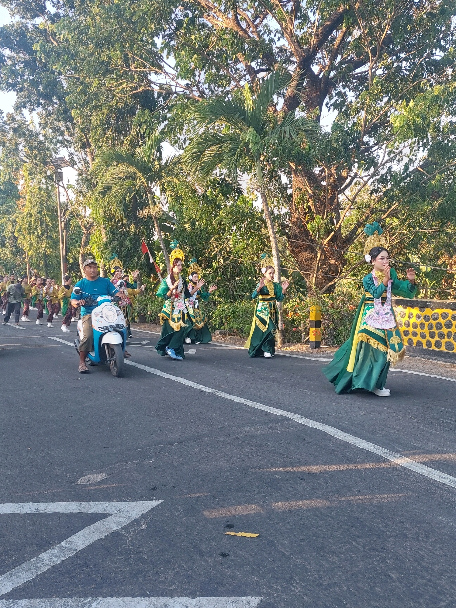 This is a medium-shot, eye-level, full-color, outdoor photograph taken on a street in Jono, Indonesia. A man wearing a blue t-shirt and shorts is riding a white and blue scooter on the right side of the road. He is looking forward and appears to be participating in a parade. Behind him, several people are walking and dancing in traditional green and gold costumes. The dancers are wearing elaborate headdresses and have their hands raised. The road is paved with asphalt and has white lane markings. Trees line the right side of the road, and a yellow barrier with black circular patterns is visible behind the dancers. The lighting suggests it is daytime, possibly late afternoon, with sunlight casting shadows on the road. The background is filled with lush green foliage and trees.