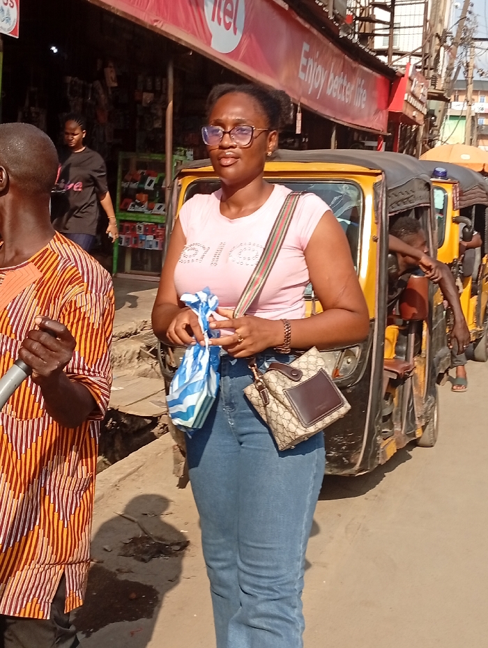 A woman wearing glasses, a pink shirt, and blue jeans stands on a sunny street. She carries a handbag and a striped plastic bag. Behind her, a yellow auto rickshaw is parked with passengers inside. To her left, a man in a patterned shirt is visible, and further back, another woman walks near a shop displaying mobile phones. An Itel sign is above the shop, and the words "Enjoy better life" are written on it.