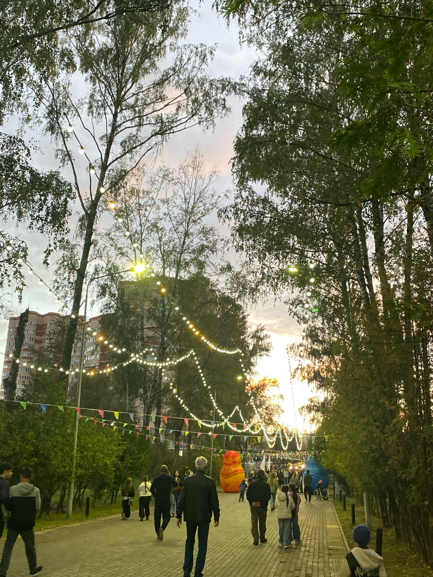 The image depicts an outdoor scene in Ivanteyevka, Russia, likely during an evening event or festival. A paved walkway is lined with trees and illuminated by strings of decorative lights and triangular flags. In the background, a large brick apartment building is visible, suggesting an urban or suburban setting. Several people are walking along the path, some in groups and others individually. Prominently featured are two large inflatable figures, one resembling a Matryoshka doll and another that is blue. The sky is overcast with hints of sunset colors. The overall atmosphere suggests a public gathering or a festive occasion.