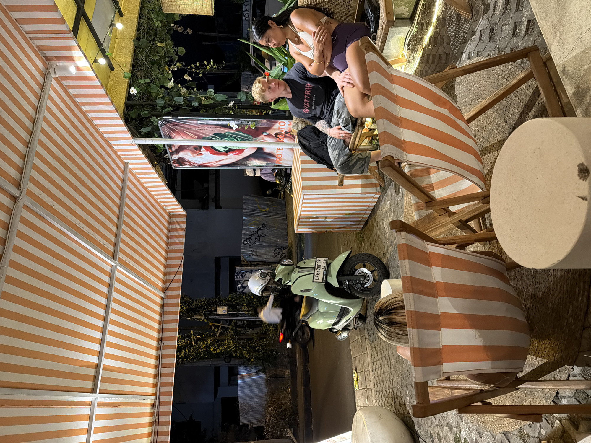 The image depicts an outdoor scene in North Kuta, Indonesia. A light green scooter with a white helmet on the seat is parked on a cobblestone surface. Adjacent to the scooter, two wooden chairs with orange and white striped fabric are positioned around a white cylindrical table. Two individuals, a man and a woman, are seated in the chairs. The man has short blond hair and is wearing a black t-shirt and camouflage pants. The woman has long dark hair and is wearing a white top and purple shorts. They appear to be in close proximity. Overhead, a striped awning provides shade. In the background, there's a banner with the word "GELATO" visible. The overall lighting suggests it is evening or nighttime.