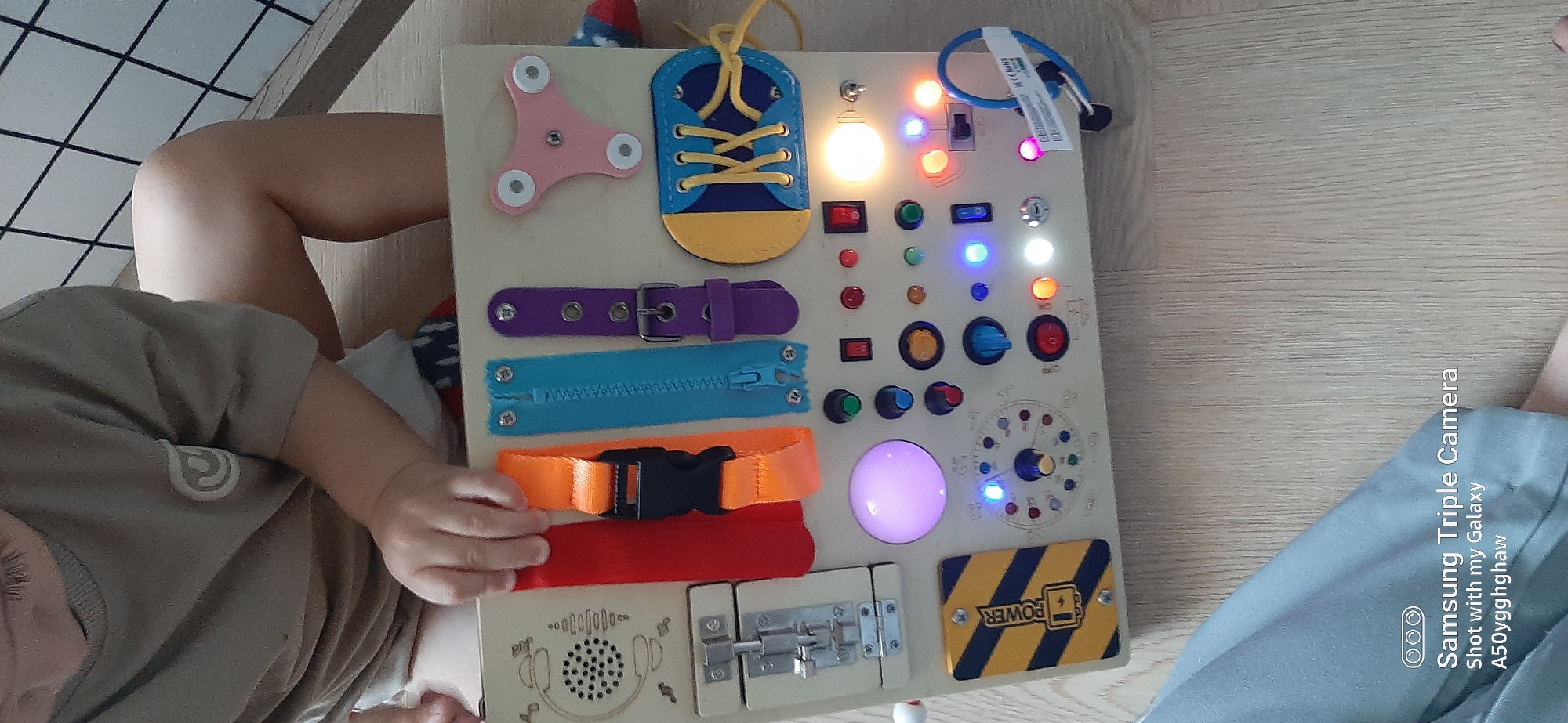A child's hands are visible on a wooden busy board with various interactive elements. The busy board is positioned on a wooden floor. Elements on the board include a red strap, an orange strap with a buckle, a blue zipper, a purple strap with a buckle, a pink spinner, a shoe with laces, a toggle switch, various colored buttons and lights, a dial with numbers and a pointer, and a yellow and black striped caution sign with "POWER" and a battery icon. Some of the lights on the busy board are illuminated. The child is wearing a light brown t-shirt and their legs are visible. The background includes a black and white checkered pattern and a wooden structure. The bottom left corner of the image contains a watermark indicating "Samsung Triple Camera" and "Shot with my Galaxy A50yggghaw". No specific location in Dubai, United Arab Emirates can be determined from the image.