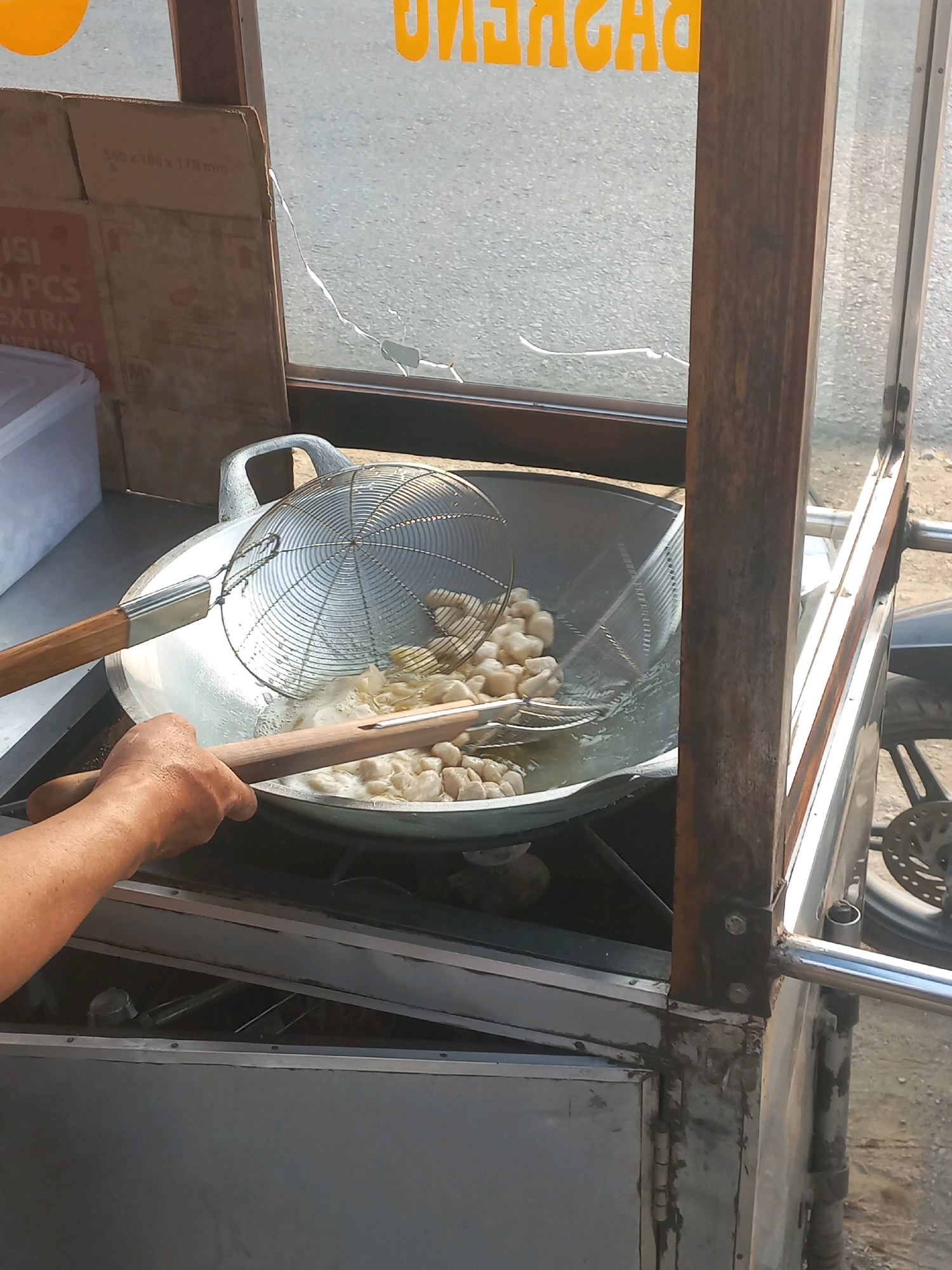 In Baturetno, Indonesia, a street food vendor is preparing food. The image captures a close-up view of a large, shallow pan filled with hot oil, where small, light-colored food items, possibly meatballs or dough balls, are being fried. A metal sieve, held by a wooden handle, is submerged in the oil, scooping up the cooked food. Another wooden utensil rests on the edge of the pan. The vendor's left hand and forearm are visible, gripping the sieve's handle. The food cart, constructed with metal and wood, is partially visible, with a clear plastic enclosure and a wooden frame. In the background, a cracked glass panel reveals a glimpse of the street and a box with text, suggesting a commercial setting. The overall scene depicts a bustling moment of food preparation in an urban environment.