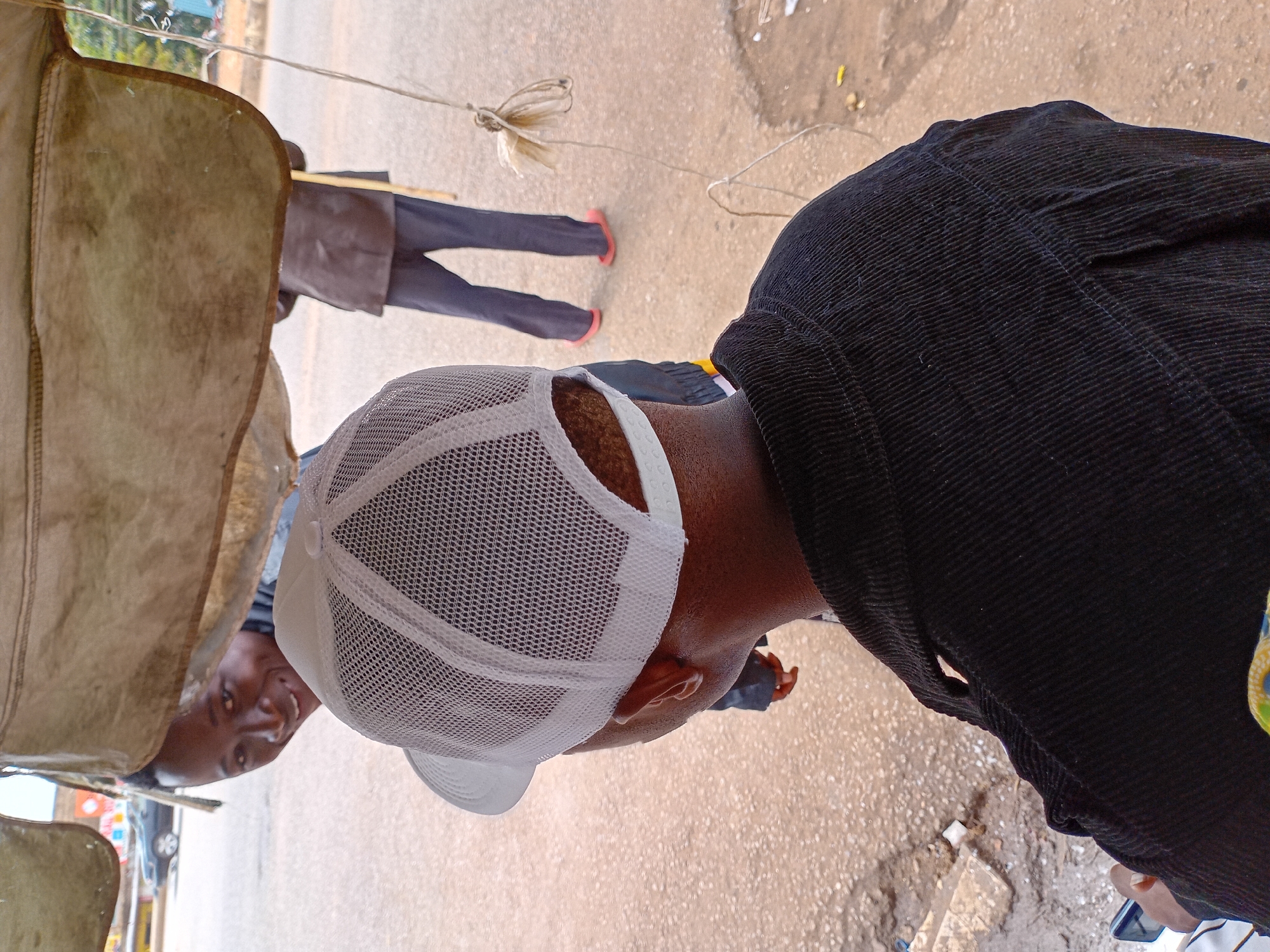 The image shows a man wearing a white mesh baseball cap and a black corduroy jacket, likely standing on a street in Bukuru, Nigeria. In the background, beneath a draped beige tarp or canvas, a second person is visible, facing the camera with a smile. A third figure is seen further in the background, wearing a brown jacket and purple pants with red shoes. A rope is visible on the ground, suggesting some form of tethering or pulling. The ground appears to be a paved road surface. The image has a slightly tilted perspective.