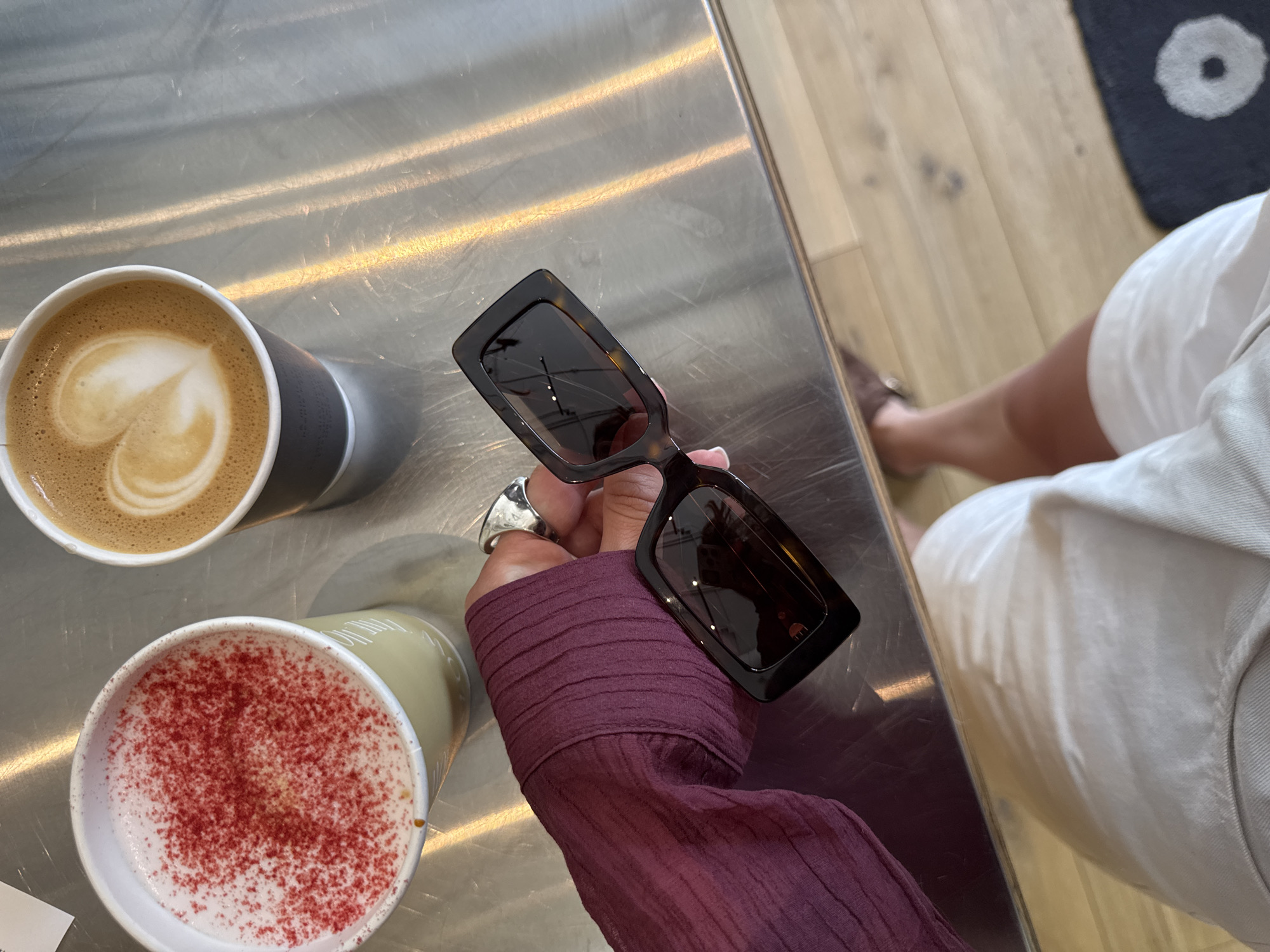 In Estosadok, Russia, a person's arm, clad in a maroon, textured sleeve, holds a pair of tortoise-shell sunglasses. Their hand, adorned with a silver ring, rests near two cups of coffee on a metallic table. One cup contains a latte with a heart-shaped foam art, while the other is topped with pink sprinkles. The scene is illuminated by streaks of light reflecting off the table's surface, casting a soft glow on the drinks and the sunglasses. A portion of the person's white shorts and legs are visible at the bottom of the frame, with a hint of a wooden floor and a dark rug in the background.