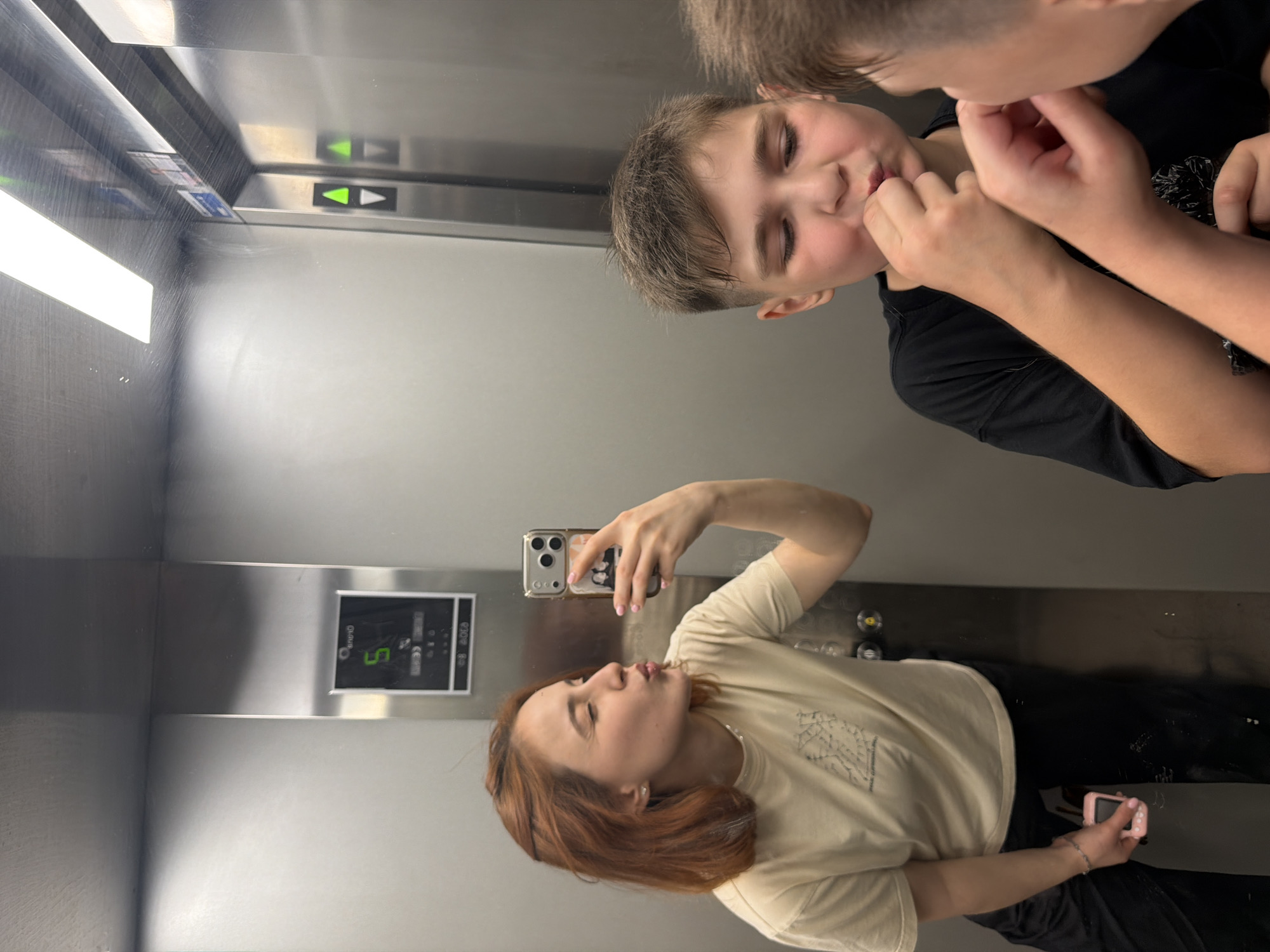 The image shows a young woman with reddish-brown hair taking a selfie in an elevator. She is wearing a light-colored t-shirt and black pants, and she is puckering her lips for the camera. In the reflection of the elevator mirror, a young boy with short brown hair is visible, also looking towards the camera and bringing his hand to his mouth. Another boy's head is partially visible in the foreground, also interacting with the first boy. The elevator interior appears to be made of stainless steel, with illuminated floor indicator lights and elevator buttons visible. The scene does not contain any elements that would suggest a location in Strasbourg, France.