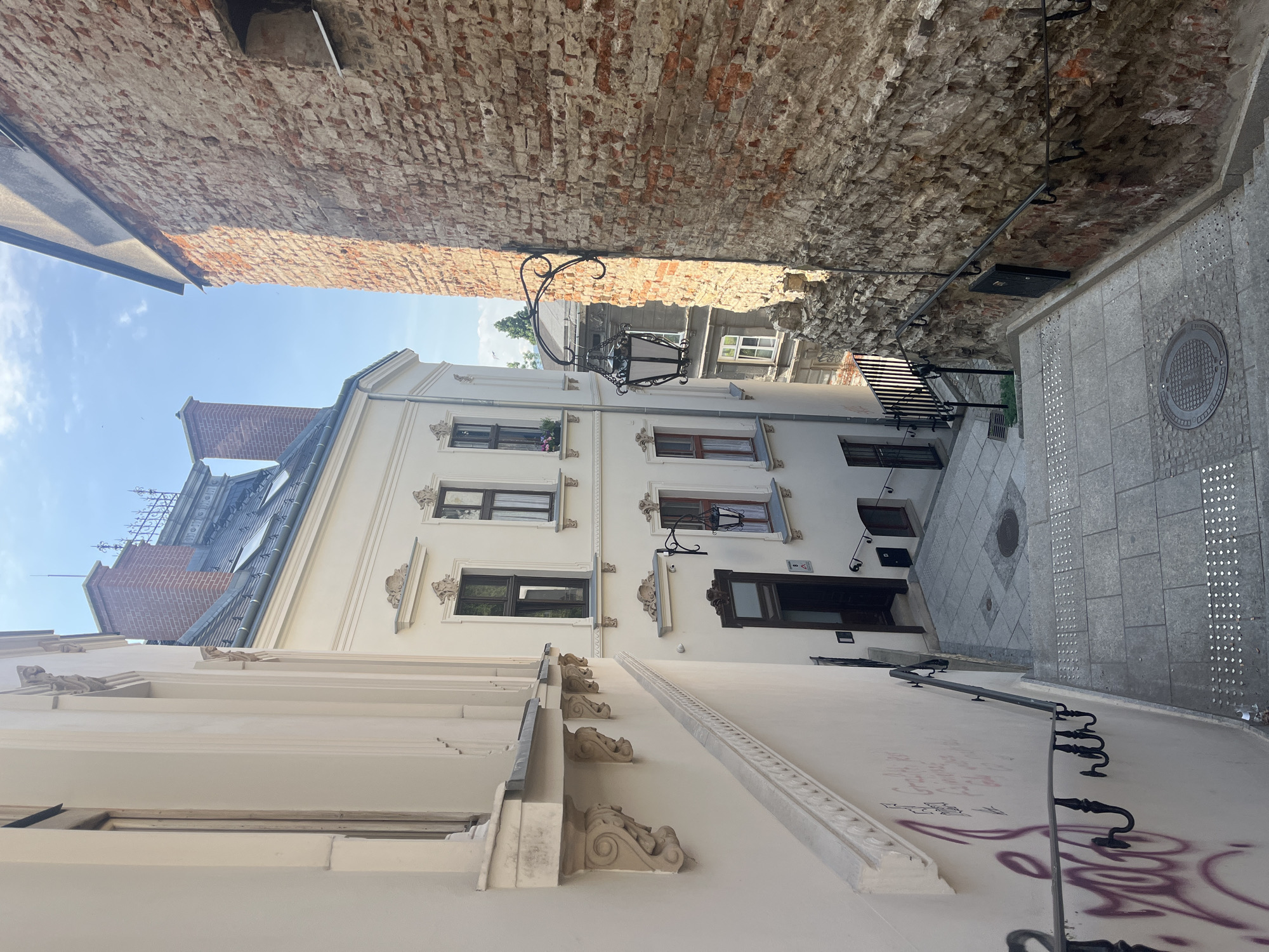The image shows a narrow cobblestone alleyway in a European city, likely Europe, with buildings on both sides. The alleyway slopes downwards and features a stone staircase with ornate railings. The building on the left is light-colored and features classical architectural details, including decorative brackets under a balcony. The building on the right is made of rough, aged brick, and an old-fashioned lantern is mounted on a bracket extending from it. The sky is blue with a few scattered clouds. The scene appears to be well-lit by natural sunlight, casting shadows that emphasize the textures of the brickwork and the steps. The alleyway has a historical feel, with a mix of architectural styles and weathered materials. The presence of graffiti on the left wall suggests a contrast between the historic architecture and modern urban elements. The overall impression is one of a charming, old-world urban environment.