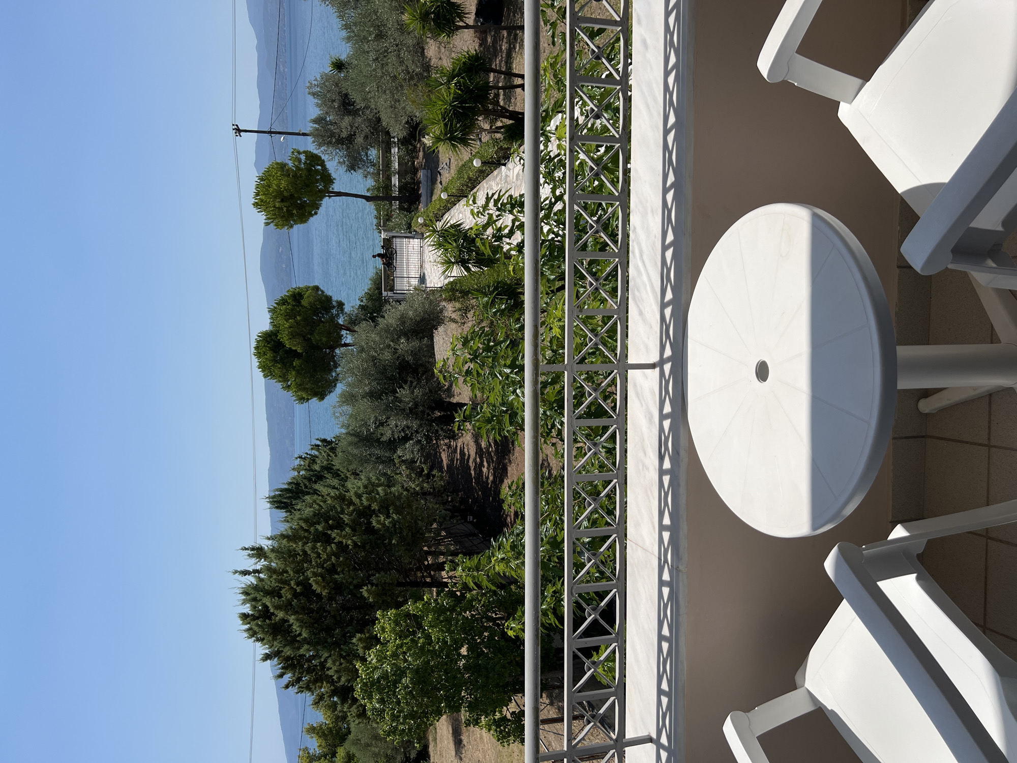The image depicts a balcony overlooking a garden and the sea. In the foreground, two white plastic chairs are positioned around a white plastic circular table. A decorative white metal railing separates the balcony from the garden. The garden features a paved walkway lined with small white globe lights, surrounded by various trees and plants. Beyond the garden, the blue sea stretches to the horizon, with a distant coastline visible under a clear blue sky. A single person on a bicycle is visible in the garden, near a gate. Power lines are visible against the sky. The overall impression is a serene outdoor setting, likely in a coastal town. This scene is located in the Municipal Unit of Eretria, Greece, a region known for its historical sites and beautiful coastline.
