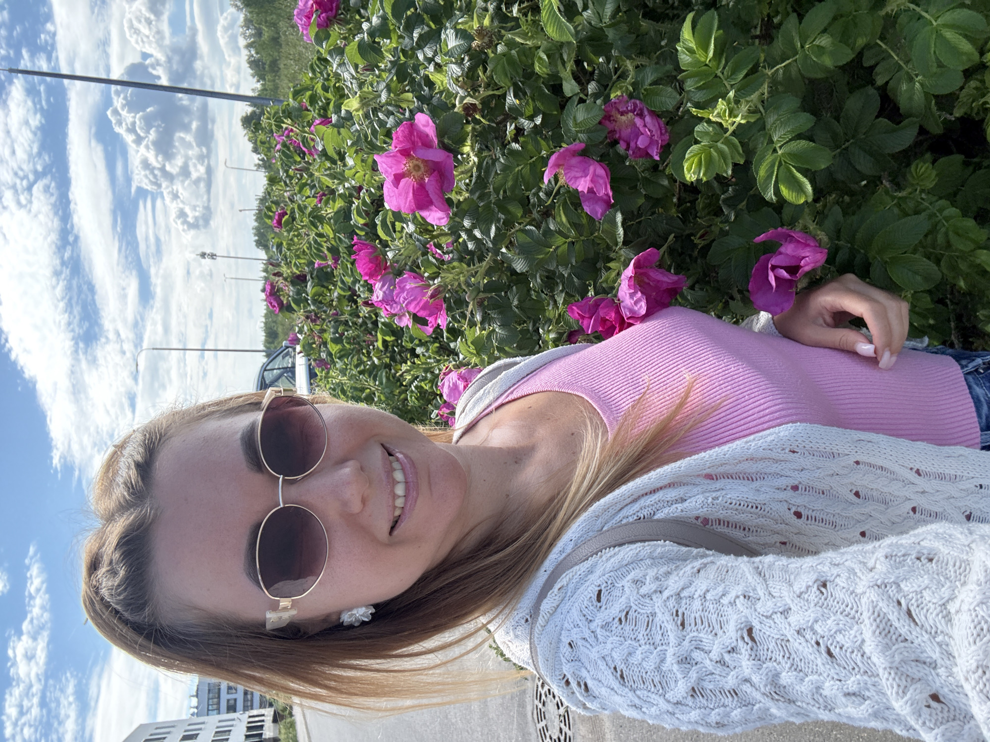 A woman stands outdoors, taking a selfie. She has blonde hair pulled back and is wearing sunglasses, a white knitted cardigan over a pink top, and blue jeans. She is smiling, and her earrings are visible. To her right, a large bush with vibrant pink flowers is in bloom. In the background, a white building, streetlights, and a cloudy blue sky are visible. The ground beneath her appears to be paved, with a circular grate.