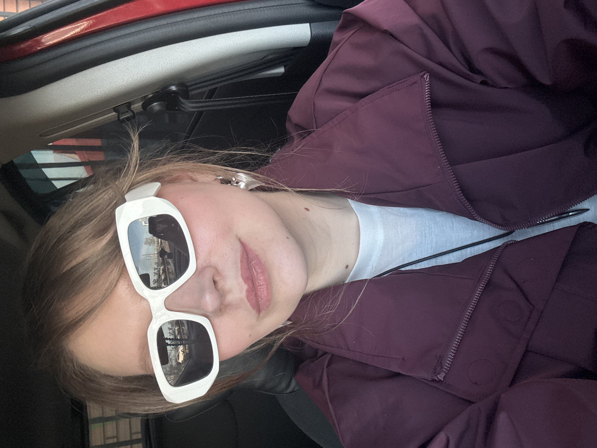 A woman wearing white sunglasses and a maroon jacket is in a car. The reflection in her sunglasses suggests she is in a parking lot. The location is Izhevsk, Russia.