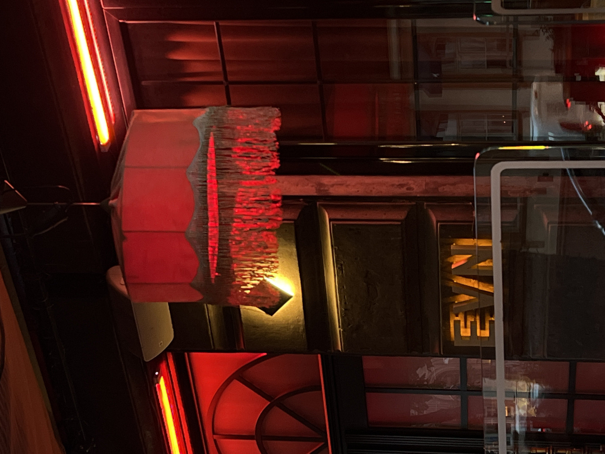 The image captures a dimly lit indoor scene in Unknown, Unknown, characterized by a prominent red color scheme. A decorative lamp with a red shade and intricate fringed trim dominates the left side of the frame. The illumination casts a reddish hue on the surrounding elements, including neon lights. The background features a grid-like pattern of dark-colored frames and red panes. Reflected in the window are letters "EVAN," in yellow. The scene's atmosphere is somewhat moody, with a mix of artificial and reflected light sources.