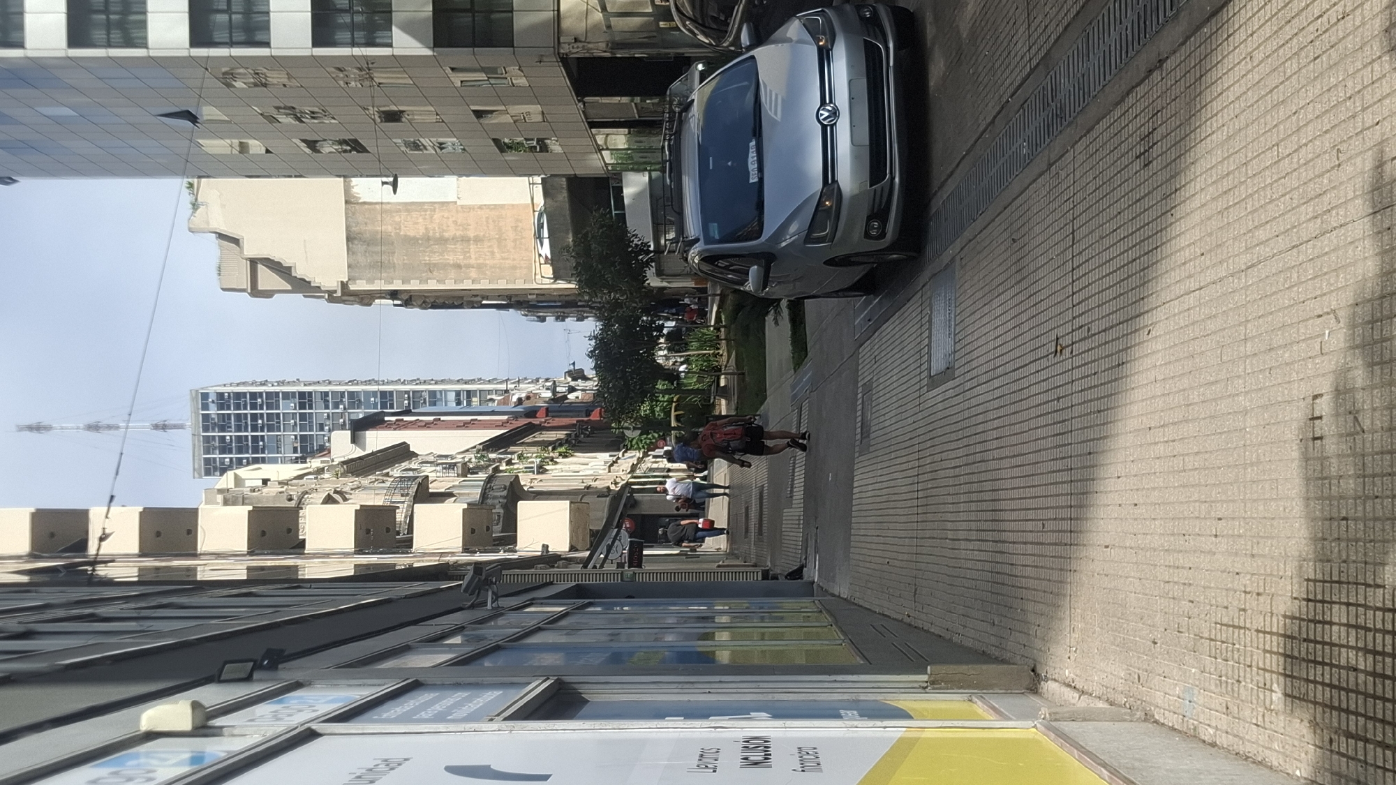 The image shows a street in Buenos Aires, Argentina. A silver Volkswagen car is parked on the right side of the street. People are walking on the sidewalk. Buildings are visible on both sides of the street. A tall building with a communications tower is in the background. The image is taken at an oblique angle.