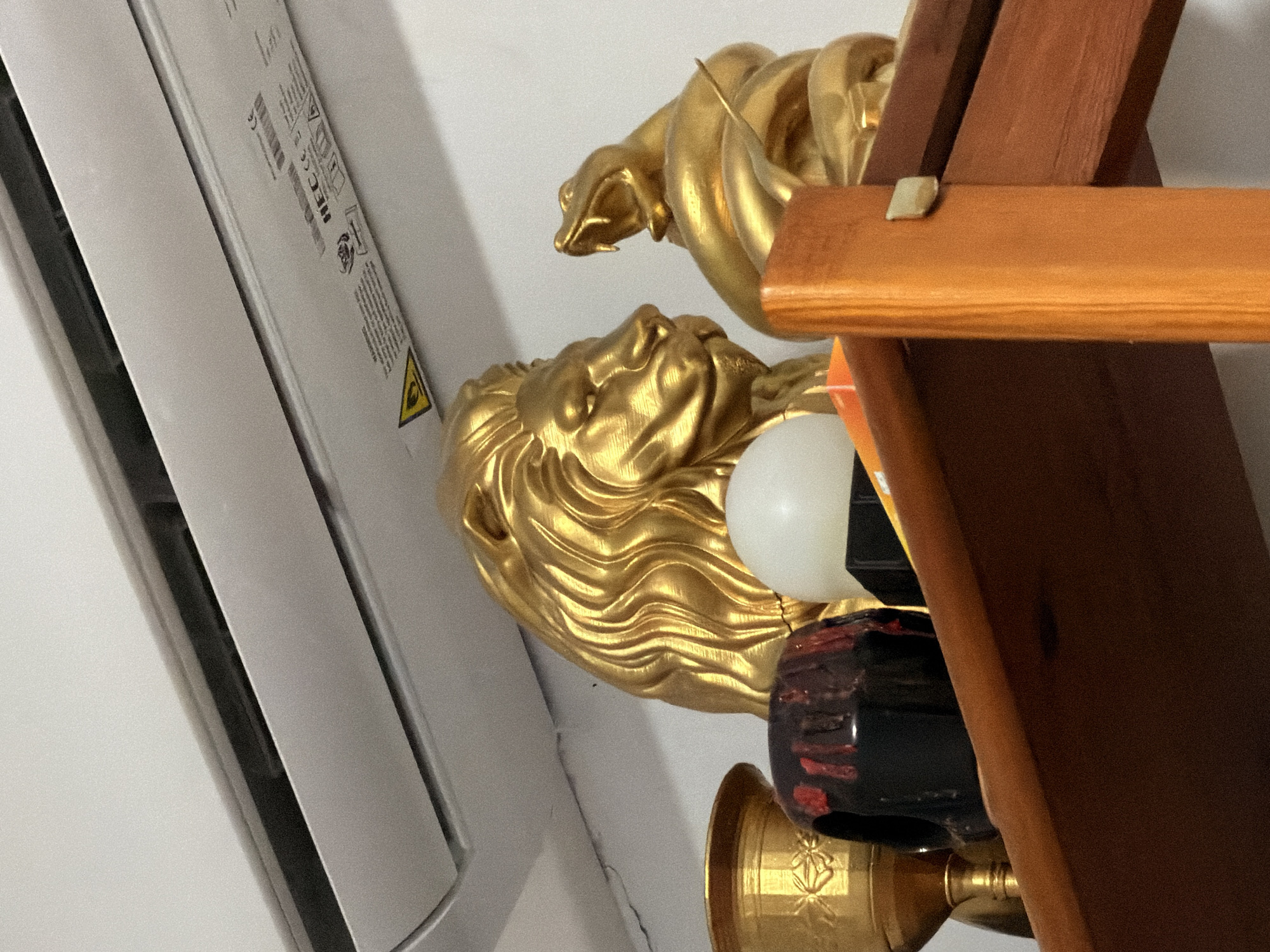 The image shows a close-up of a shelf with decorative items. On the shelf are two golden statues, one of a lion's head and the other of a coiled snake with its mouth open. To the left of the lion's head is a golden chalice with intricate carvings. Next to the chalice is a black ceramic object with red markings, resembling a stylized skull or pot. In front of the lion statue, there is a small white spherical object, possibly a light or a decorative orb. The shelf itself appears to be made of wood. Above the shelf, a white air conditioning unit is mounted on a white wall. The overall impression is that of a curated display of decorative pieces on a shelf in a room. No specific location in Torremolinos, Spain, can be determined from the visual information provided.
