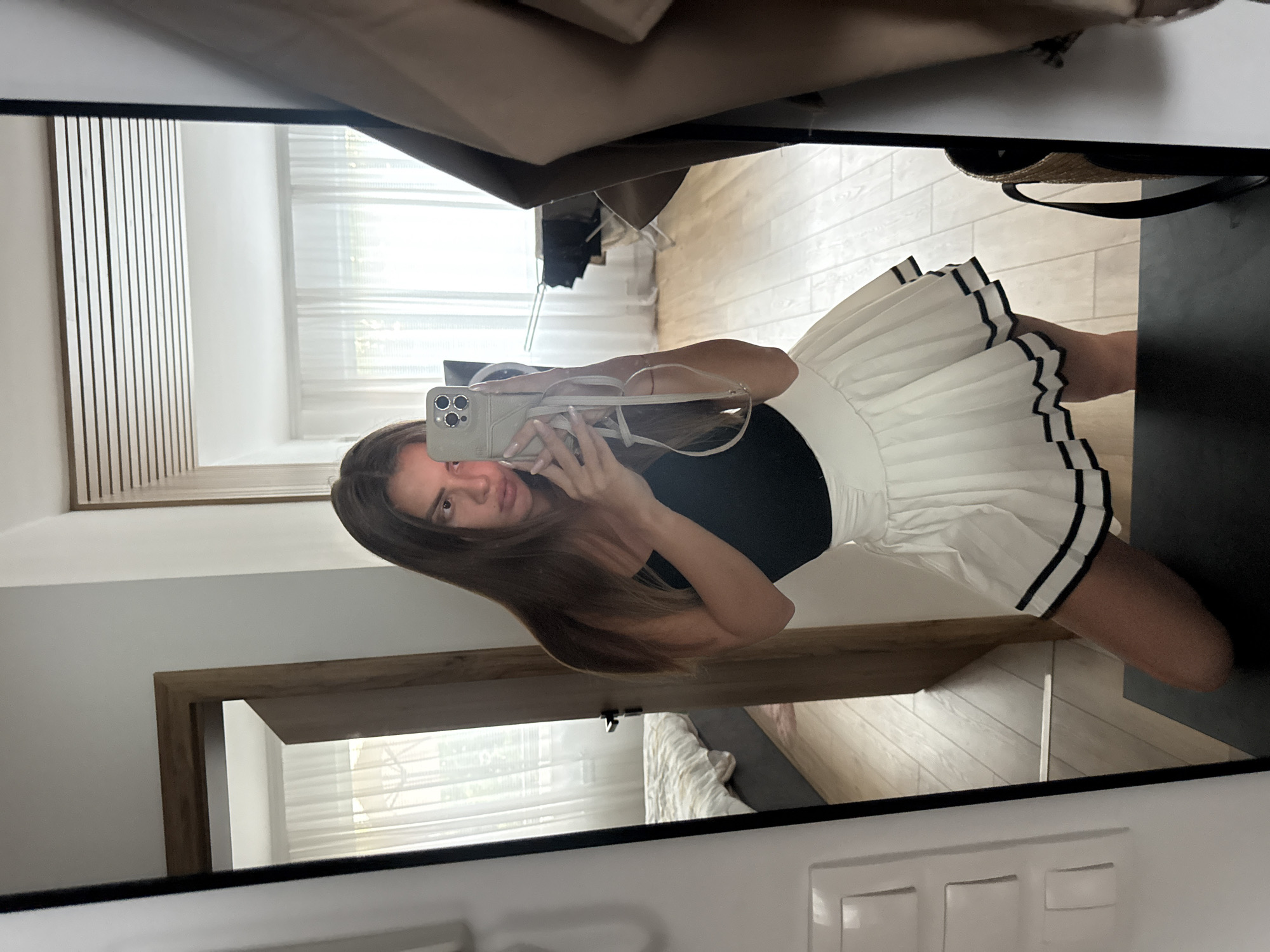A young woman, reflected in a full-length mirror, poses for a selfie. She is wearing a black tank top and a white pleated mini-skirt with black trim. Her right hand holds a smartphone, capturing the image. The mirror reflects a room with light-colored walls and wooden flooring. In the background, visible through an open doorway to the left, is a bed with white and patterned bedding. To the right of the woman, a tan jacket hangs on a hook. The scene appears to be an indoor residential setting, possibly an apartment or house. The location is not specified as being in Plovdiv, Bulgaria.
