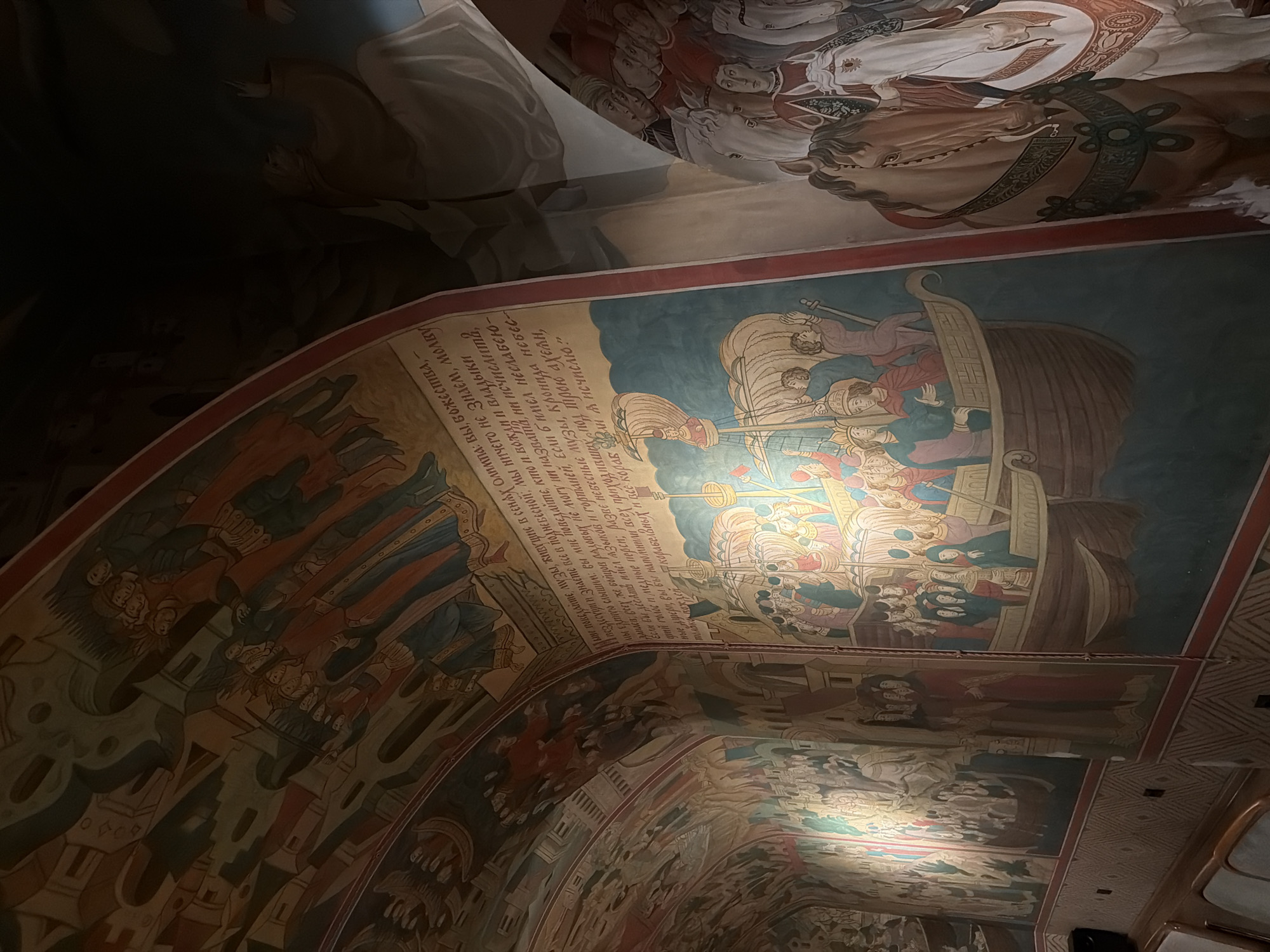 This media file captures a ceiling fresco inside a building, likely a church or historical site, given the style of the artwork. The fresco depicts a narrative scene with numerous figures, rendered in a traditional religious or historical style with vibrant colors. The main panel visible shows a group of people on a boat, engaged in an activity that seems to be a journey or a significant event. The figures are depicted with distinct clothing and expressions, suggesting a story unfolding.
Above this central panel, another section of the fresco shows figures in ceremonial attire, with a prominent depiction of a horse and rider, possibly signifying royalty or a religious leader. The artwork extends across the curved ceiling, with multiple panels and detailed illustrations that suggest a larger, overarching narrative. Some figures appear to be in procession, while others are engaged in different actions, all contributing to a complex visual story.
Visible text in a Cyrillic script is present within one of the fresco panels, possibly accompanying the imagery with narration or identifying the characters and events. The text appears to be an invocation or a historical account. The overall impression is one of grandeur and historical significance, with the artwork creating an immersive and awe-inspiring atmosphere. The lighting in the image suggests an indoor setting, possibly dimly lit to preserve the ancient artwork. The style of the painting and the subject matter point towards a location with a rich cultural and religious heritage, consistent with the provided context of Moscow, Russia.