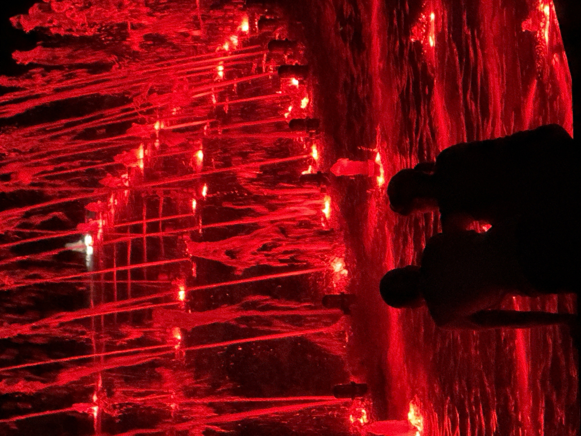A dimly lit, close-up, vertical, outdoor shot depicts a fountain illuminated by red lights at night. In the foreground, the silhouetted backs of two people are visible, one facing left and the other facing right. They are seated, with their heads and shoulders prominent. The water of the fountain is agitated and reflects the red light. In the background, streams of water are illuminated in red as they fall, creating a dramatic effect. Rows of small lights are visible beneath the falling water, adding to the visual spectacle. The overall impression is one of a vibrant and dynamic water feature.