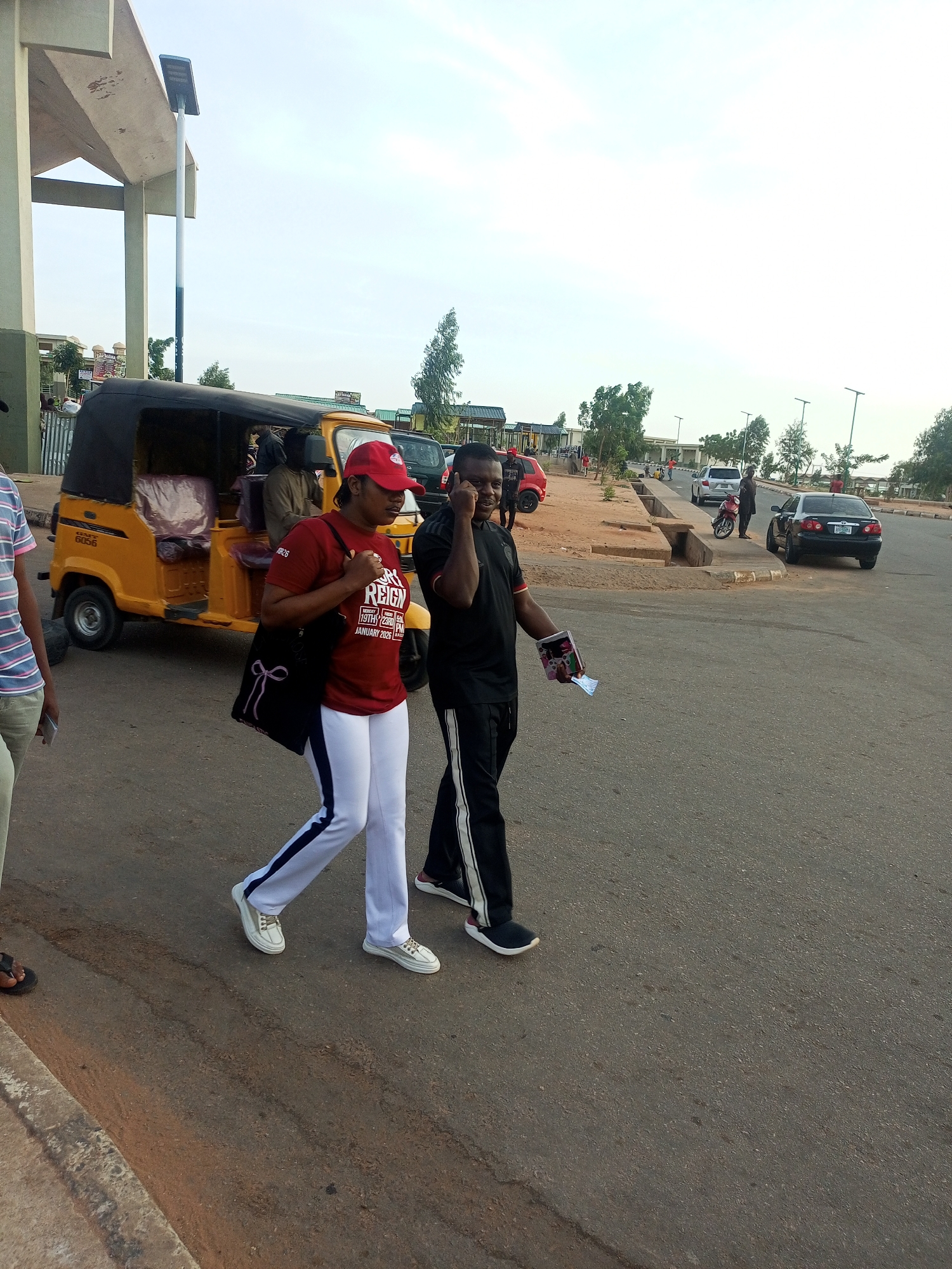 The image captures a street scene in Gombe, Nigeria. A woman wearing a red t-shirt, red cap, and white pants with a dark stripe walks alongside a man in a black track suit, who is on a phone call and holding some papers. They are walking on an asphalt road. In the background, there is a yellow auto rickshaw (tuk-tuk) with passengers inside. Several cars, including a black sedan, are parked or driving in the distance. The environment includes sidewalks, buildings, trees, and streetlights. The sky is overcast.