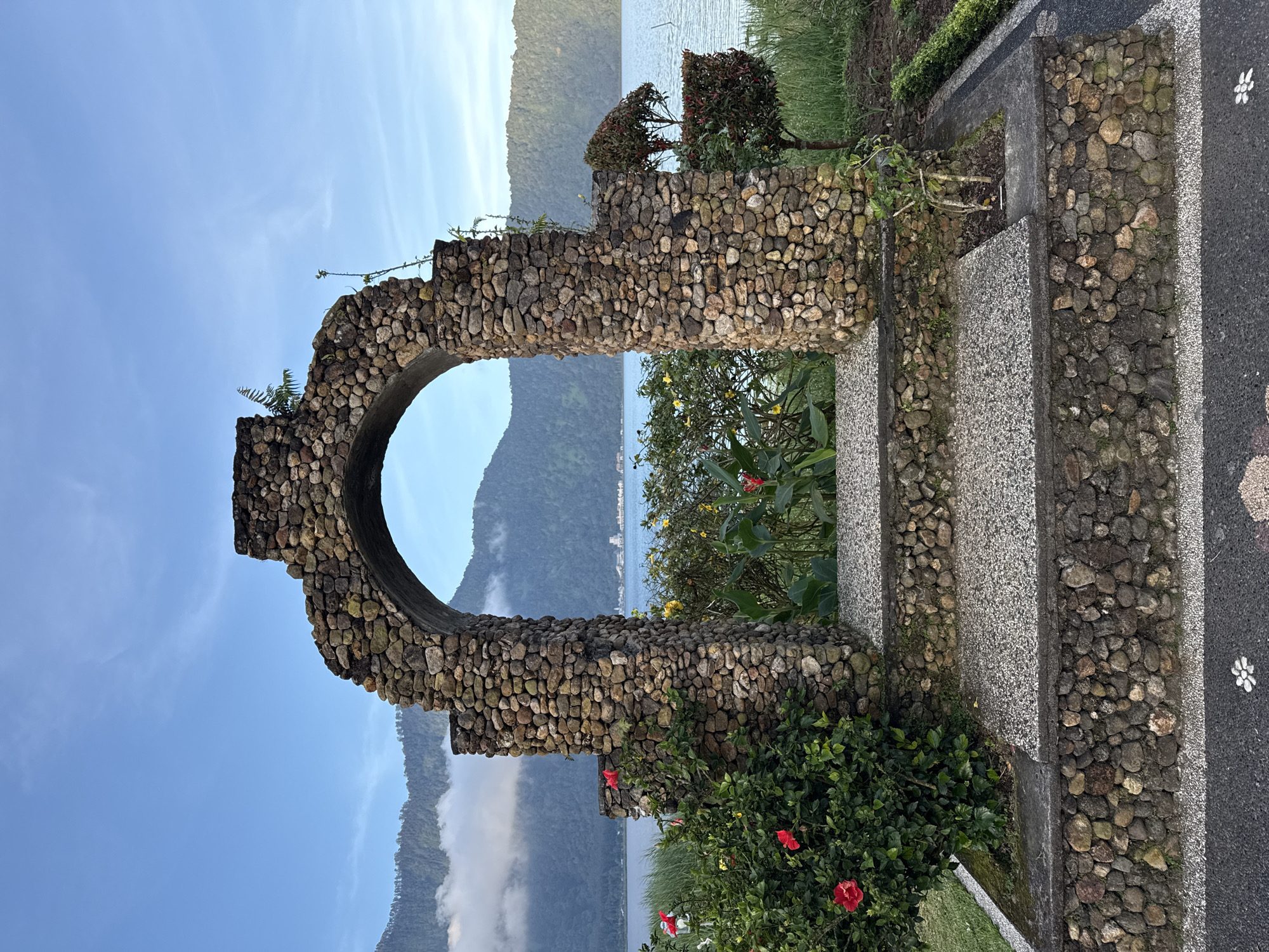 The image captures a stone archway and a set of steps made of stone and concrete, seemingly part of a garden or park feature. The archway is constructed from small, rounded stones, creating a rustic and weathered appearance. Through the archway, there is a view of a lake or ocean, with mountains visible in the background. The sky is blue with some clouds. The steps lead up to the archway and are also constructed with stone and concrete. Green vegetation and flowering plants surround the structure, adding a touch of nature to the scene. The photograph was taken in Baturiti, Indonesia.