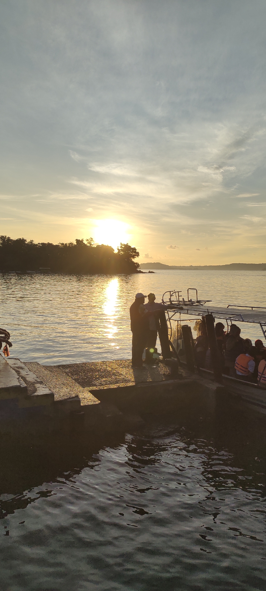 This is a vertical, medium-shot, outdoor, daytime photograph taken at sunset, likely on the water's edge near the city of Sabang, Indonesia. The sun is low on the horizon, casting a warm, golden glow across the scene and reflecting on the water. The sky is a mix of blue and wispy white clouds, with the sun's rays breaking through.
In the foreground, a concrete pier or dock extends into the water. On the edge of the pier, two men are standing. One man, wearing a light-colored shirt and a dark cap, is facing the water. The other man, in a darker shirt and a dark cap, is standing next to him and pointing towards something off-camera. They appear to be interacting.
To the right of the men, a covered boat is docked. The boat has a canopy and appears to be carrying several passengers, some of whom are wearing life vests, visible in orange. The boat is positioned alongside the pier, suggesting it is either about to depart or has just arrived.
In the middle ground, across the water, is a tree-covered island or landmass. The trees are silhouetted against the bright sun and its reflection on the water. In the background, the horizon stretches across the frame, with more distant land visible. The water is relatively calm, with ripples reflecting the sunlight. The overall atmosphere is peaceful and serene, characteristic of a sunset by the sea.
