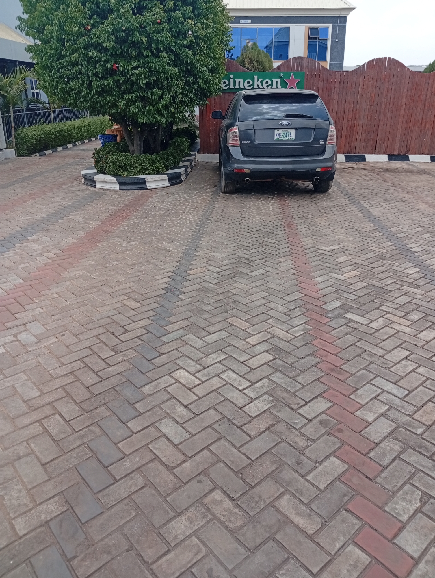 The image shows a gray Ford Edge SUV parked in an outdoor area paved with gray and red bricks arranged in a herringbone pattern. Behind the car is a wooden fence and a building with blue windows, above which a "Heineken" sign is affixed. To the left of the car, there is a tree surrounded by a round planter bordered with black and white stripes. The scene appears to be in Adankolo, Nigeria, as indicated by the vehicle's license plate. The sky is overcast. There are no people visible in the frame.