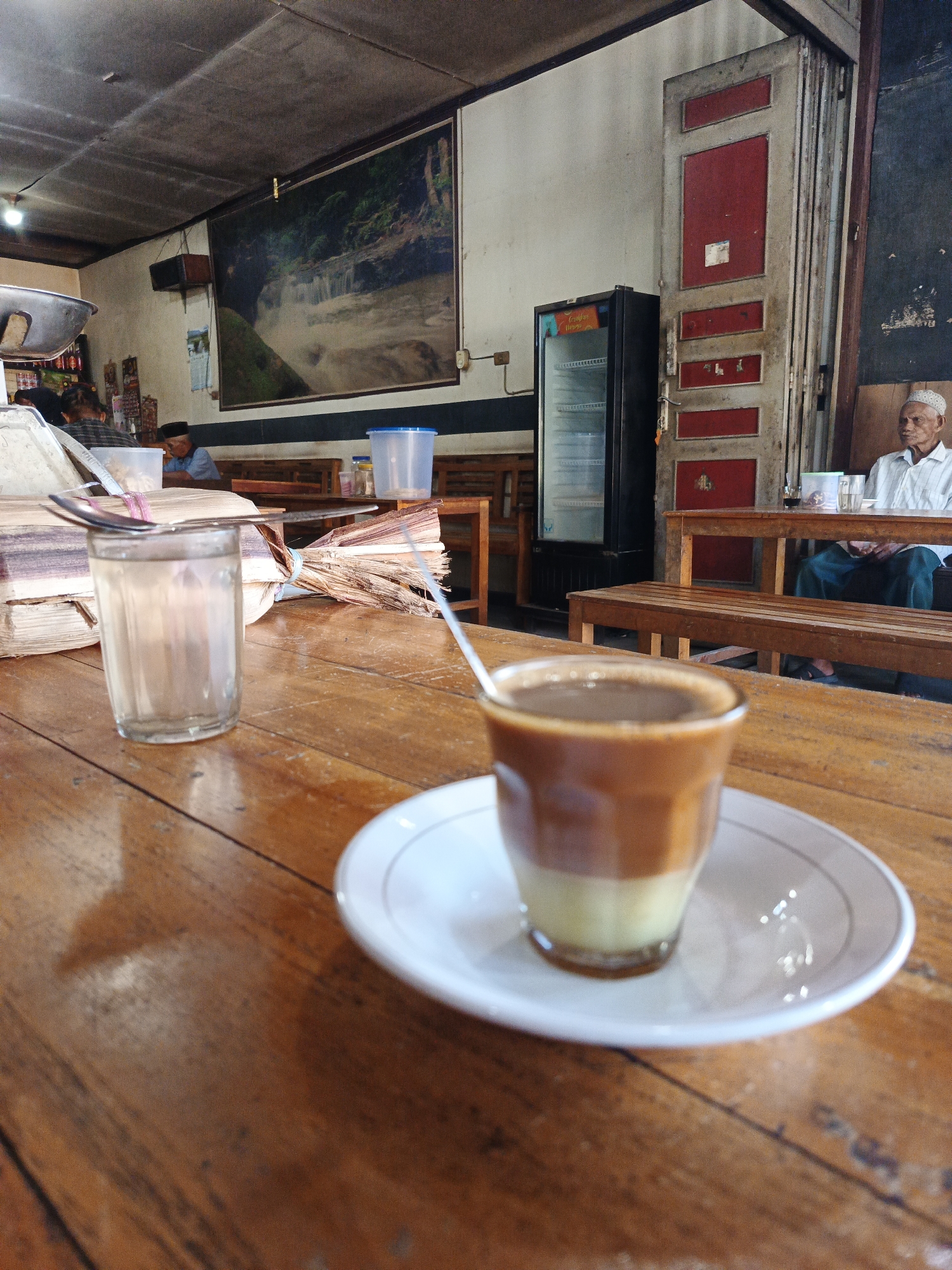 The image depicts the interior of a cafe, likely in Geulumpangmeujim-Jim, Indonesia. Focus is on a wooden table with a cup of coffee on a saucer and a glass of water, and a spoon, along with bundled palm sugar. In the background, there are wooden benches and tables occupied by people. An elderly man is sitting at a table in the background, wearing a white shirt and a white cap. A black refrigerator is visible to his left, and a red door beside it. A large painting hangs on the wall. The ceiling is visible at the top of the frame. The overall lighting is subdued.