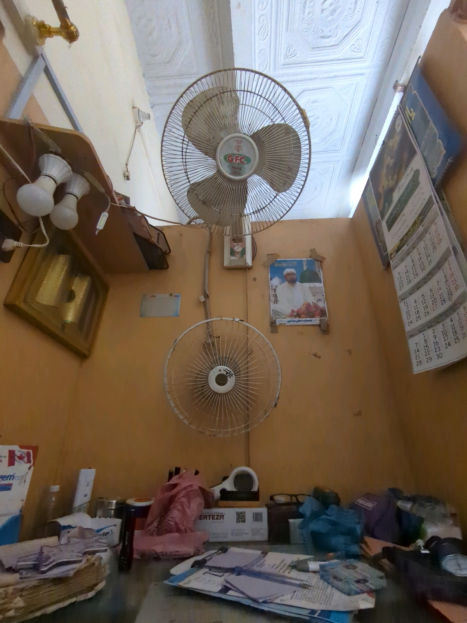 The image captures a cluttered room, likely in Karachi, Pakistan, featuring two electric fans mounted on a pale orange wall. The larger fan is a ceiling fan, and a smaller oscillating fan is mounted lower on the wall. Several items are visible on shelves and a table: a framed picture, calendars, light bulbs, and various papers and bags. The ceiling is decorated with patterned panels. The overall impression is one of a functional but untidy space.