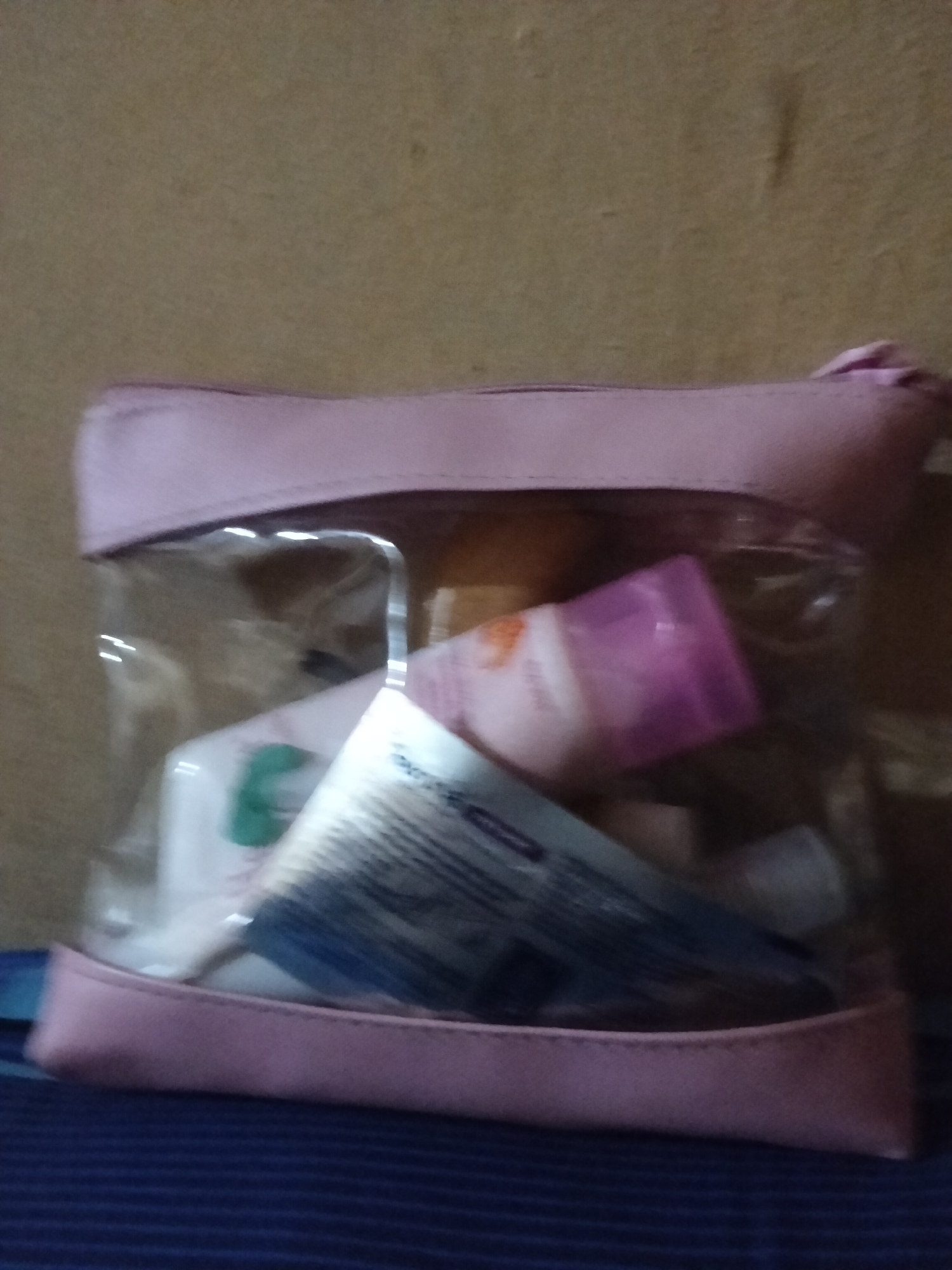 The image shows a close-up, slightly blurry view of a transparent pink cosmetic bag. The bag is filled with various personal care items, including what appear to be tubes of lotion or cream and a comb with a blue and white handle. One tube has a pink cap and a white body with orange text and a light orange graphic. Another tube is mostly white with some green visible. The bag is made of clear plastic with pink trim and a pink zipper. The background is a textured, light brown surface, possibly a wall. The bag is resting on a surface with dark blue and lighter blue stripes, suggesting it might be on a bed or a couch. The lighting is dim, and there are some reflections on the clear plastic of the bag. No people are visible. The scene appears to be indoors. There is no clear indication of the time of day or weather. No text is legible within the image.