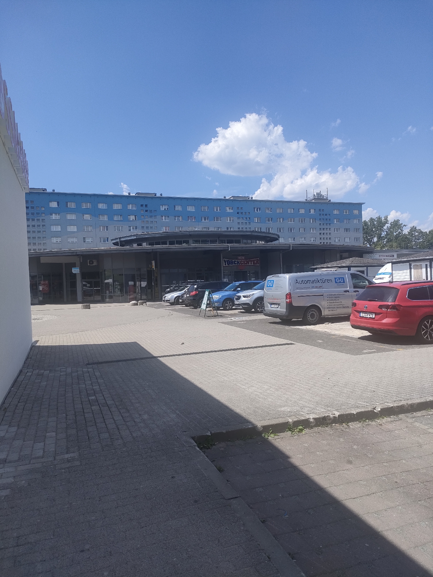 This media file captures an outdoor scene on a sunny day, likely in Chemnitz, Germany. The prominent subject is a light blue, multi-story building that appears to be an apartment complex or office building, with many uniformly spaced windows. In front of this building is a commercial area with glass-fronted shops, one of which has a "SALE %" sign visible.
Several vehicles are parked in the lot in front of the shops, including a silver van with "Automatiktüren GU" (Automatic Doors GU) and other text in German printed on its side, a red station wagon, and a blue SUV. A white truck is also partially visible. The ground is paved with interlocking bricks and concrete. The sky is a bright blue with scattered white clouds, suggesting fair weather and daytime. The overall impression is a mundane, everyday scene in a commercial or residential area. No people are clearly visible.