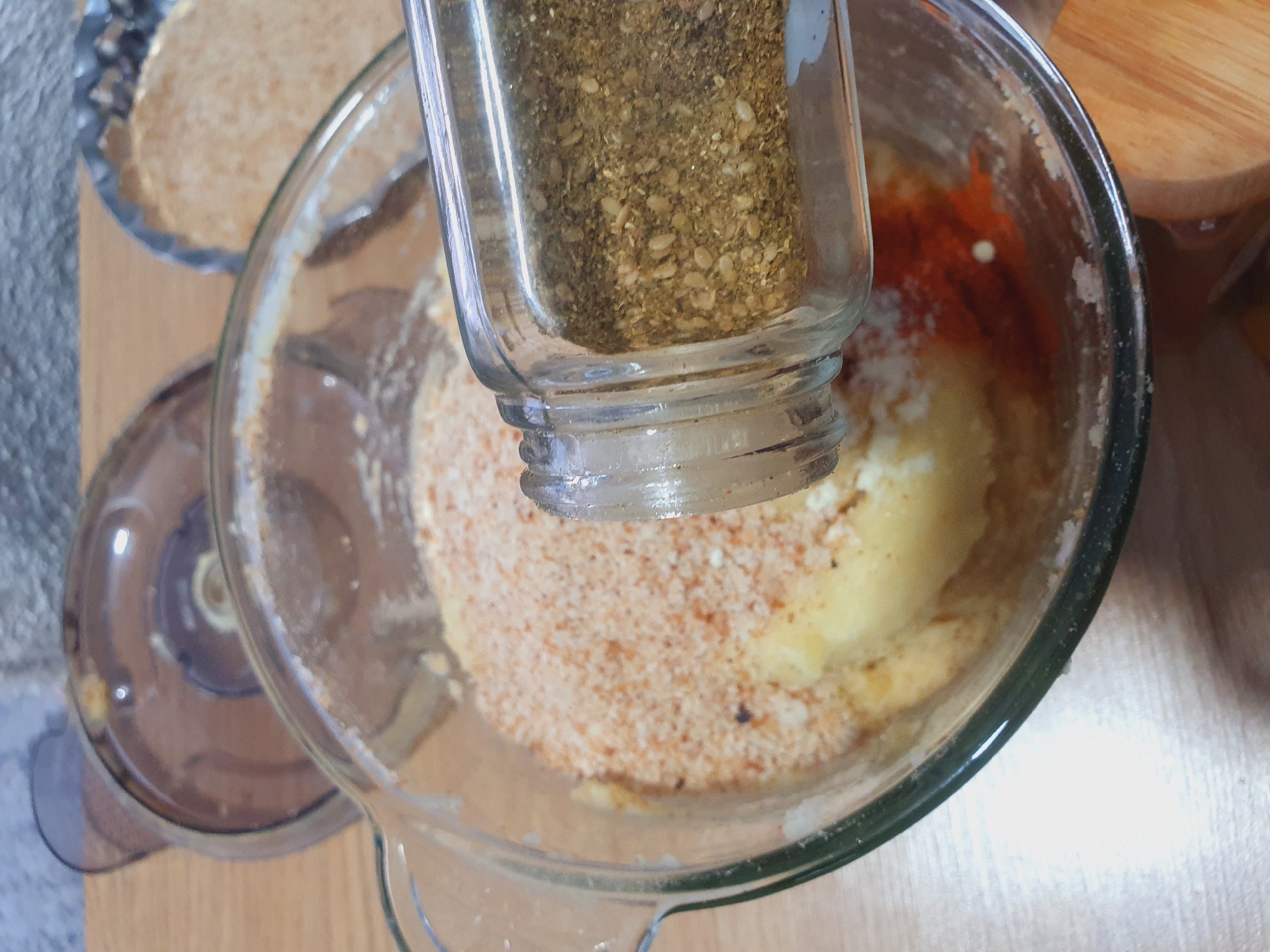 A close-up shot captures a glass bowl filled with a mixture of ingredients, including what appears to be mashed potatoes, breadcrumbs, and red spices. A glass spice jar, tilted at an angle, is pouring more spice into the bowl, its contents a blend of small seeds and dried herbs. The scene is set on a wooden surface, with blurred background elements suggesting a kitchen environment, possibly including a food processor lid and another dish containing breadcrumbs. The lighting is bright, highlighting the textures and colors of the food ingredients.