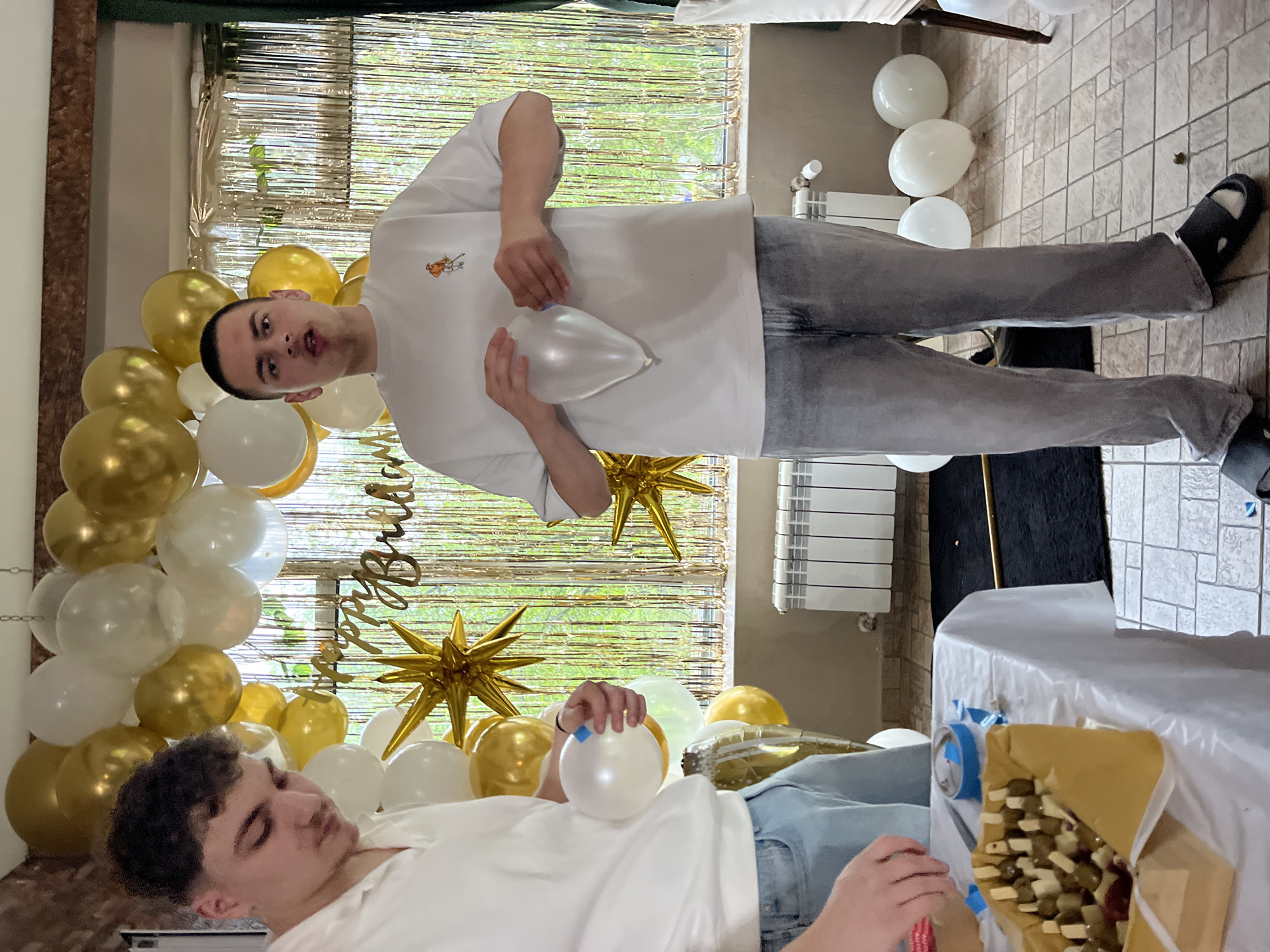 Two people are shown indoors in front of a balloon and star decoration. One person stands wearing a white shirt and grey jeans, holding a balloon. The second person lays on a table covered with a white tablecloth, wearing a white shirt and light blue jeans. There are snacks visible on the table, along with other decoratives. There is a window behind the balloon decoration. The location is unknown.