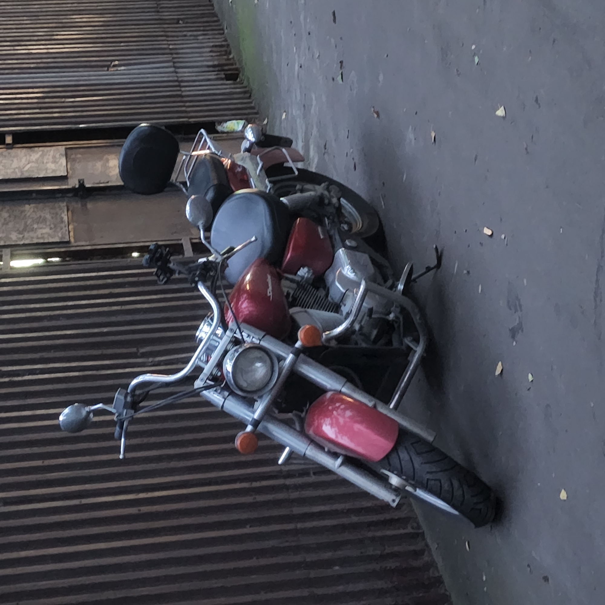The image shows a red motorcycle parked next to a vertical corrugated metal fence. The motorcycle has a black seat and a chrome exhaust pipe. The ground is paved with asphalt and there are a few fallen leaves scattered around. The lighting suggests it is either early morning or late afternoon. The fence and the overall aesthetic of the scene suggest an urban environment, possibly a back alley or service area within the city of Moscow, Russia. There are no people or animals visible in the image.