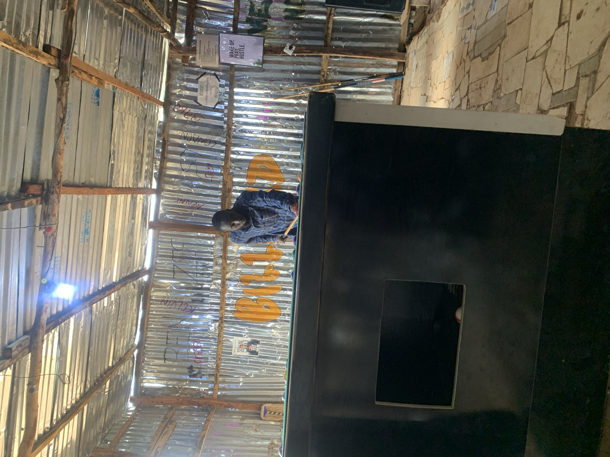 In the city of Durumi, Nigeria, a man is playing pool inside a structure with corrugated metal walls and a tin roof. He is wearing a blue plaid shirt and is positioned behind a pool table, holding a cue stick. The pool table has green felt and several colored balls visible. The walls are adorned with graffiti and posters, including one that reads "WAKE UP. PRAY. HUSTLE." There are also several pool cues leaning against the wall. The floor is paved with irregular stone tiles. The overall environment appears to be a casual, possibly open-air, establishment.
