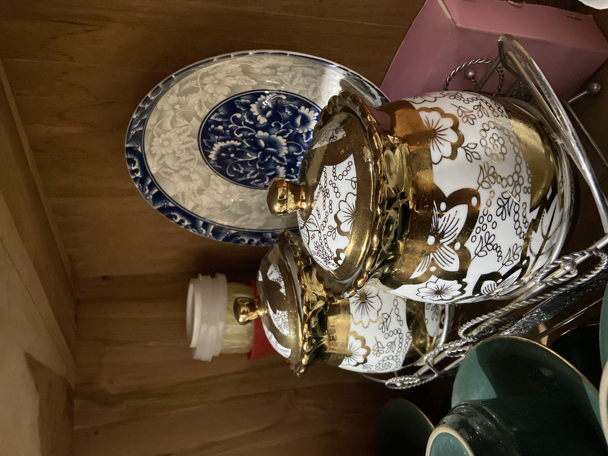A close-up, eye-level shot shows a collection of vintage porcelain items displayed on a wooden shelf. In the foreground, two lidded canisters with ornate gold floral and vine patterns on a white background are prominently featured. They are placed on a silver metal rack with twisted handles. To the left, a stack of green ceramic cups and saucers, with gold trim, are partially visible. Behind the canisters, a large, decorative plate with intricate blue and white floral patterns stands upright. The background consists of the wooden shelves and a glimpse of a pink box on the right. The lighting is dim, creating shadows and highlighting the reflective surfaces of the gold and silver. The scene suggests an indoor display, likely a home or shop, showcasing decorative dishware. There are no people or animals present in the image. The image does not contain any information about the location in Margorejo, Indonesia.