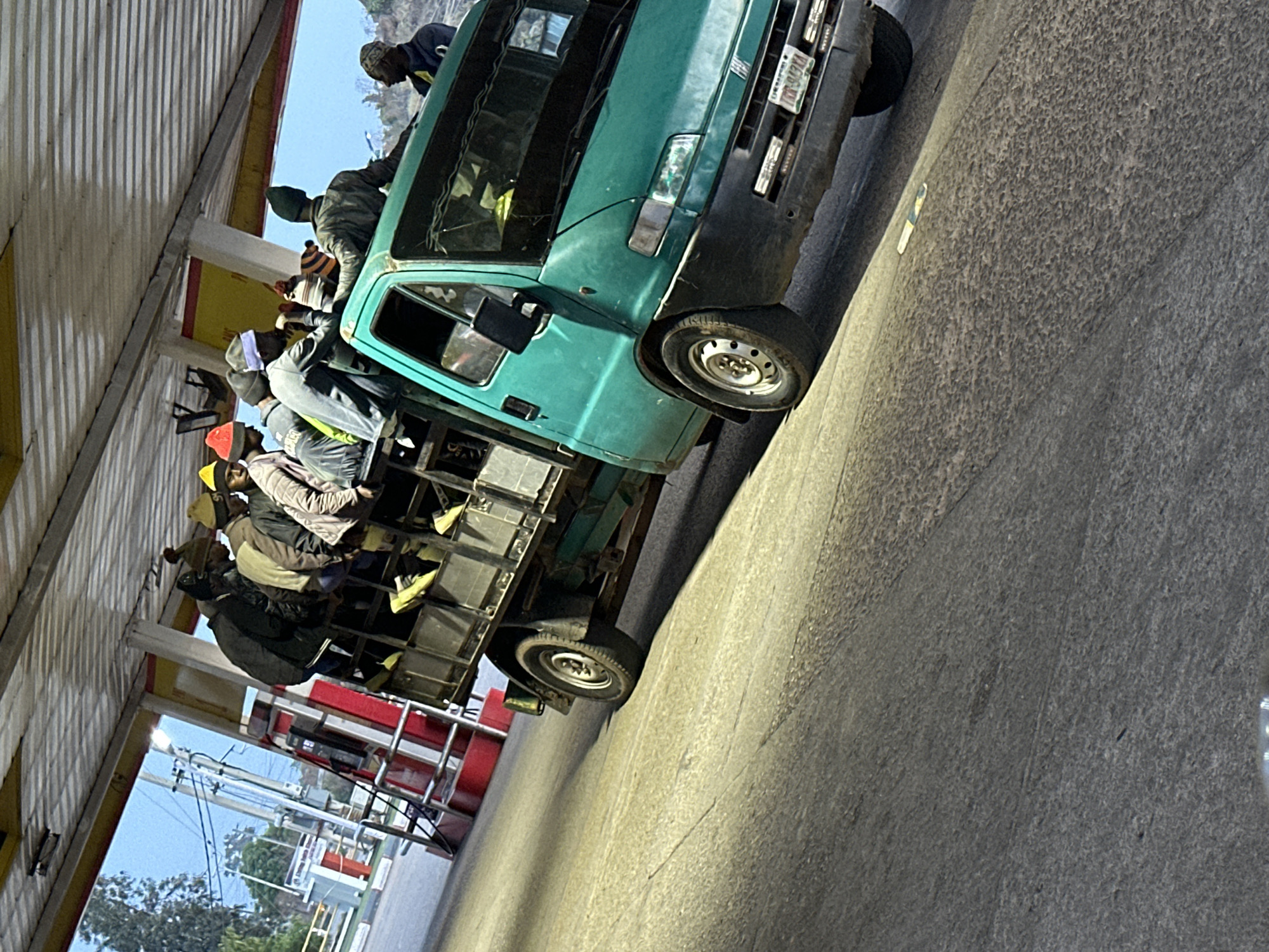 The image, taken in Gura Top, Nigeria, depicts a teal-colored pick-up truck heavily loaded with numerous passengers in its open cargo bed. The truck, which appears to be a light utility or flatbed model, is functioning as a shared public transport vehicle. Many individuals, predominantly men, are visible, some standing, others sitting on the built-in metallic framework and side rails of the truck bed. They wear various casual clothes, with several sporting hats or beanies.
The vehicle displays a license plate on its front bumper that clearly shows "LAGOS" and the alphanumeric sequence "KVN-942HA", indicating its registration in Lagos, Nigeria. The truck itself shows signs of frequent use.
The scene is set outdoors, under a structure with a corrugated metal roof and supportive pillars, which could be part of a market, a transport stop, or a roadside shelter. The ground beneath the truck is paved. In the background, a clear blue sky is visible above distant trees and utility poles, suggesting it is daytime, possibly late afternoon or early morning due to the directional lighting. The image captures a common form of informal, crowded transportation.