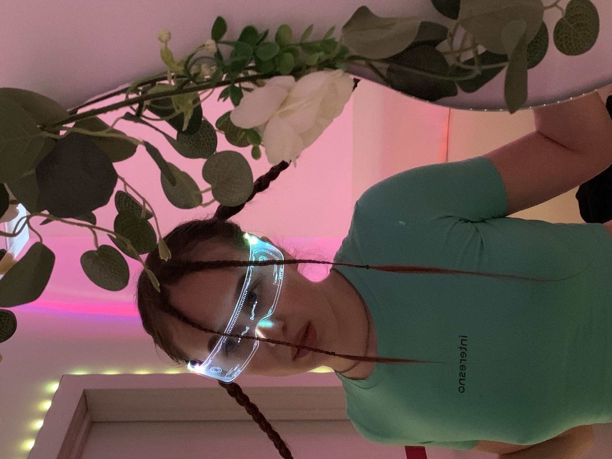 A woman wearing illuminated futuristic glasses and a light green shirt is shown in an indoor scene. The lighting has a pink and purple hue, possibly from LED lights. There are artificial leaves and a white rose arranged around her head as if in a halo. The location is unknown, the city of Фоки, Russia, is speculation.