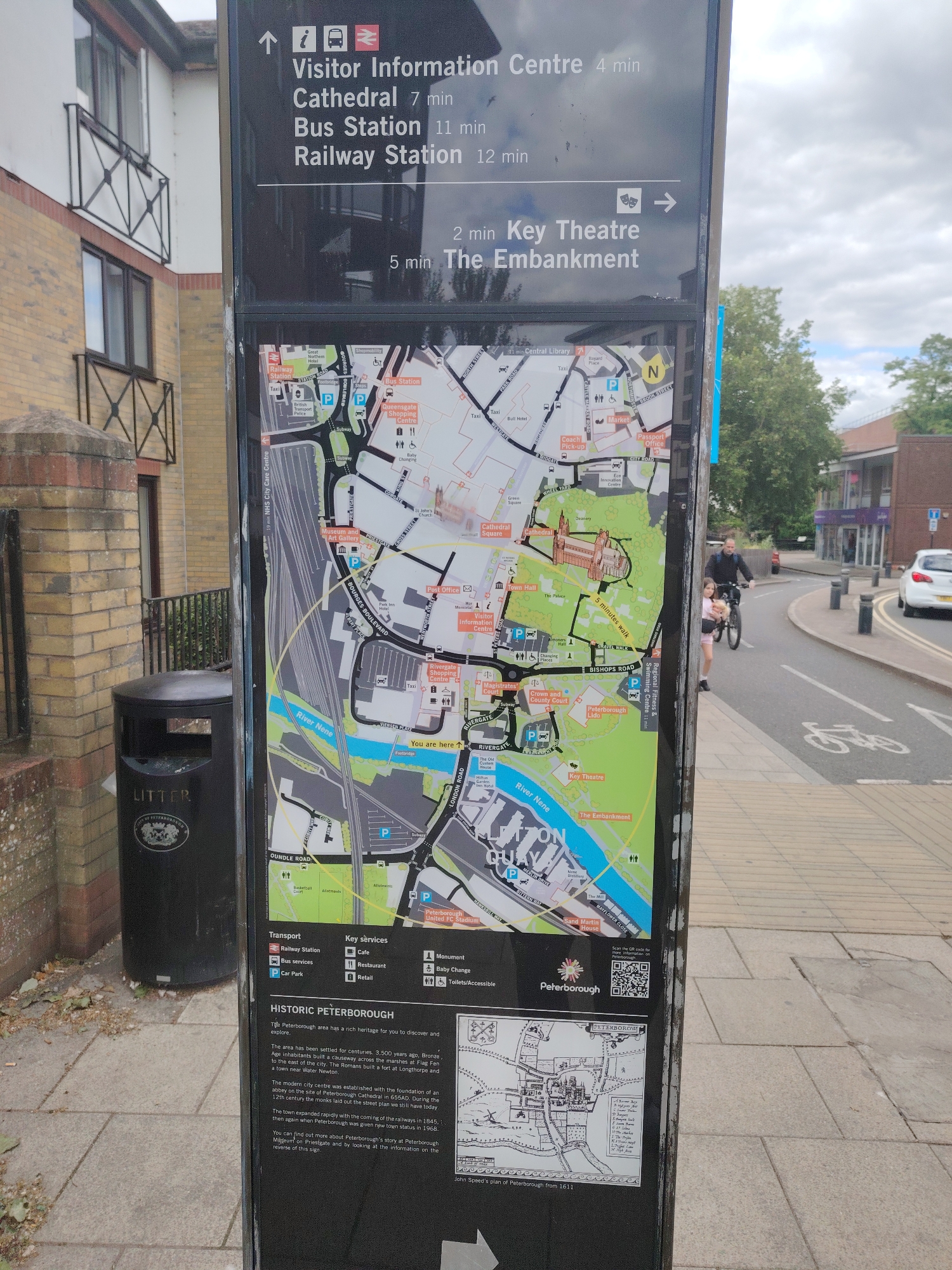 A tall, dark information sign stands prominently in the foreground, displaying a map of Peterborough, United Kingdom, along with text directions and historical information. The sign indicates walking times to destinations such as the Visitor Information Centre (4 min), Cathedral (7 min), Bus Station (11 min), Railway Station (12 min), Key Theatre (2 min), and The Embankment (5 min). The map highlights the "You are here" location near Rivergate, adjacent to the River Nene, and details various landmarks including the Cathedral, Queensgate Shopping Centre, Museum and Art Gallery, and Peterborough Lido. A legend identifies symbols for transport services (Railway Station, Bus services, Car Park) and key services (Cafe, Restaurant, Retail, Monument, Baby Change, Toilets/Accessible). A section titled "HISTORIC PETERBOROUGH" provides a brief history of the city. A QR code for more information about Peterborough and an image of "John Speed's plan of Peterborough from 1611" are also visible.
To the left of the sign, a light brown brick building with multiple windows and dark metal balcony railings is partially visible. A black litter bin with "LITTER" embossed on it is situated next to a brick pillar at the base of the building. To the right of the sign, a paved pedestrian area transitions into a road. A male individual on a bicycle, wearing a dark jacket, is visible on the road, accompanied by a female individual (possibly a child) in a pink top and shorts walking beside him. A white car is parked further down the road, and trees and other urban buildings are visible under a cloudy sky in the background. Road markings for a bicycle lane are discernible on the asphalt.