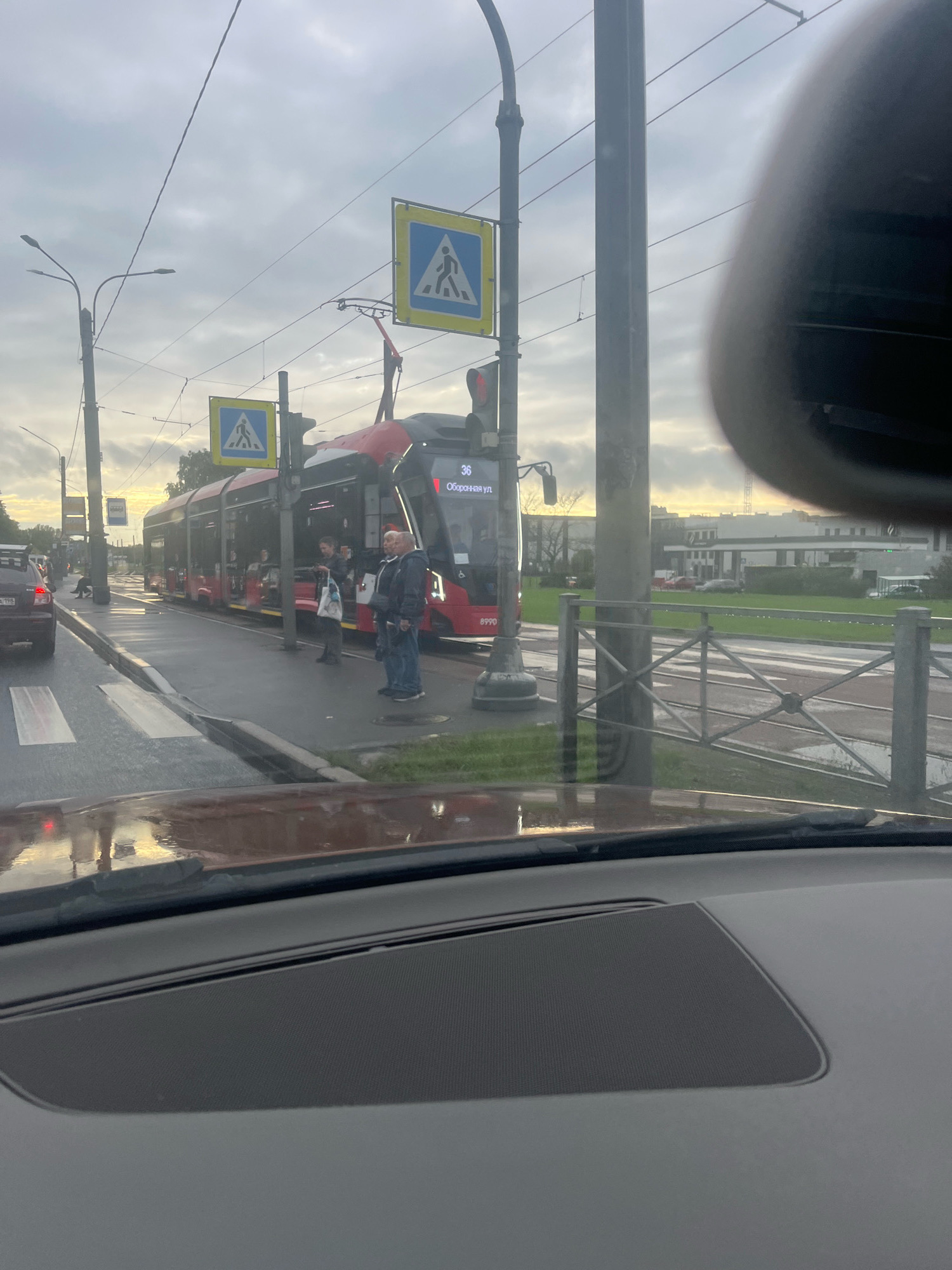 The image depicts a street scene in Saint Petersburg, Russia, likely on a cloudy or overcast day. In the foreground, the interior of a car is visible, with a portion of the dashboard and windshield. The road appears wet, suggesting recent rain.
Dominating the mid-ground is a long, red tram, numbered 36, stopped at a designated stop. The tram is modern in design, with large windows. Several people are visible on the platform, waiting to board or disembark. Two men stand near the front of the tram, engaged in conversation, while others are further down the platform. A pedestrian crossing sign is prominently displayed near the tram stop.
To the left of the tram, traffic is visible, including a dark-colored car. Streetlights and overhead wires for the tram system are present. In the background, buildings and some greenery can be seen. The overall impression is of a typical urban public transportation scene.