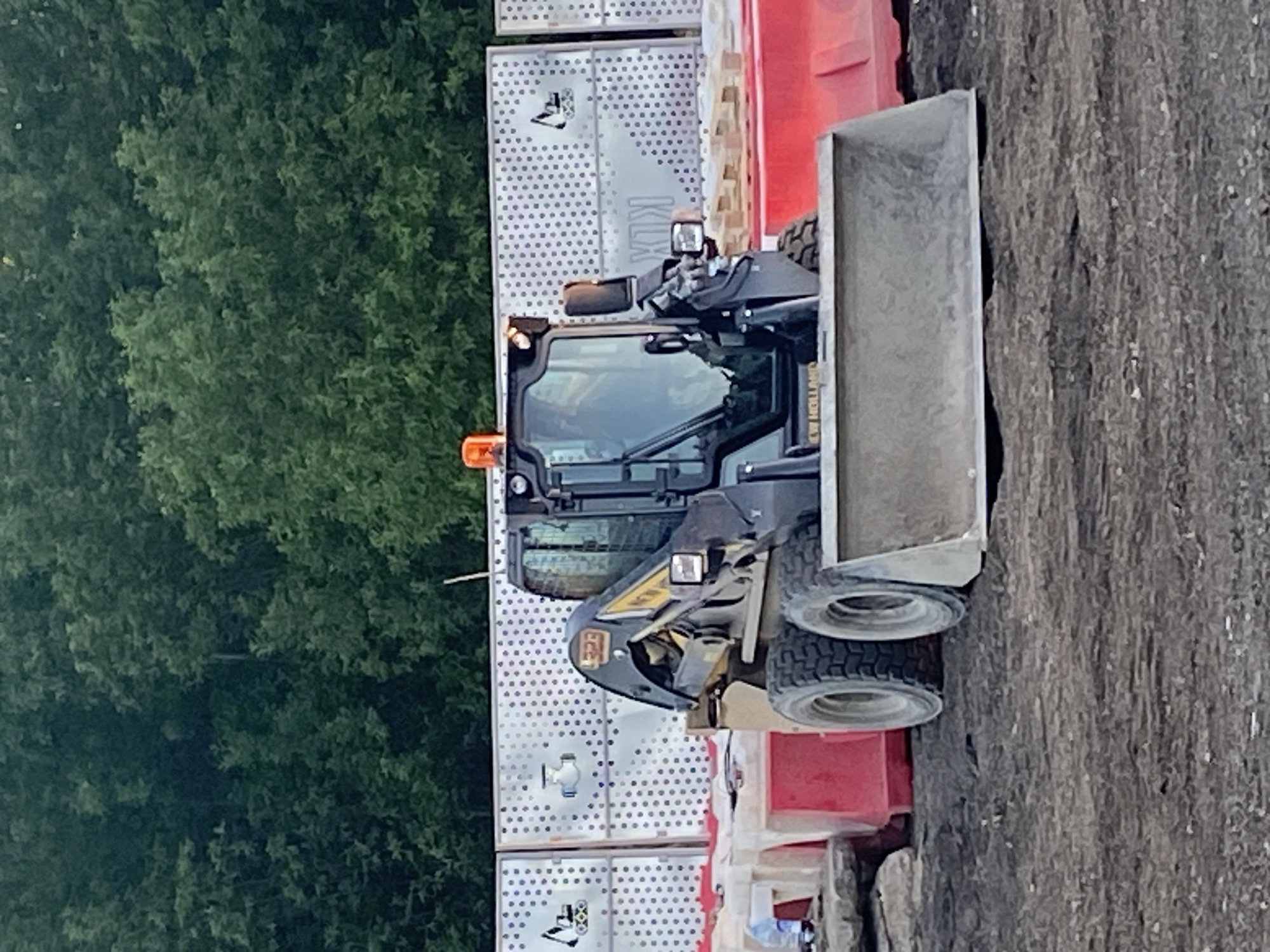 A construction site scene shows a skid-steer loader. The loader is equipped with a bucket attachment. The background includes a dense forest and metal barriers. The location is not discernible as Moscow, Russia, based on the image alone.