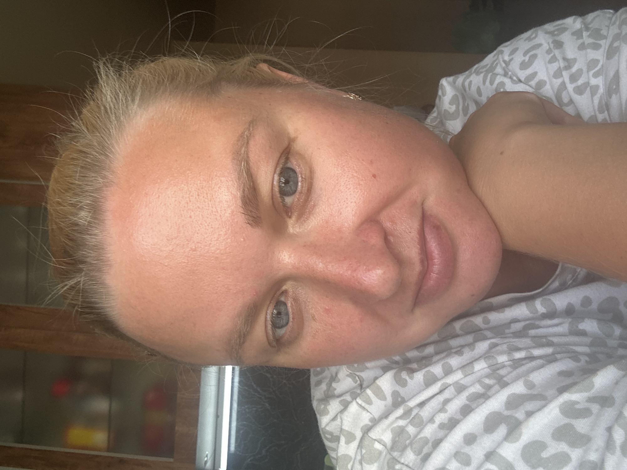 The image is a close-up, head-and-shoulders selfie of a woman with blonde hair and blue eyes, resting her chin on her hand. She is wearing a gray and white patterned shirt. The background appears to be an indoor setting with wooden cabinetry and shelving visible. The lighting suggests it is daytime. The woman's pose and facial expression are relaxed. There are no identifiable landmarks suggesting a specific location in Chaikovsky, Russia.
