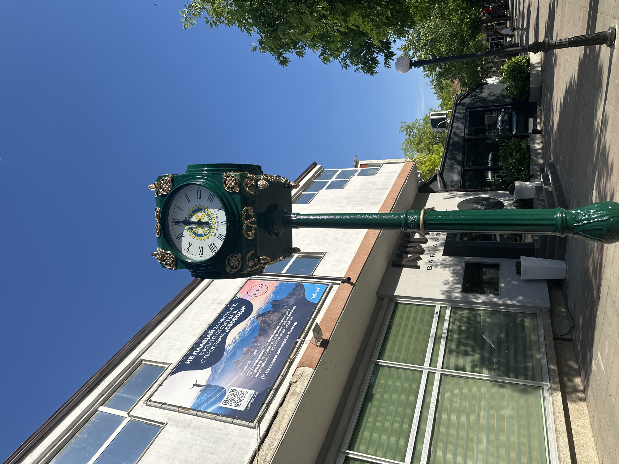 Here is a detailed description of the image:
**Scene:** The image captures a sunny street scene in Montana, Bulgaria, featuring a distinctive green clock post in the foreground. The clock has a white face with Roman numerals and a Rotary International emblem in the center.
**Environment:** The backdrop consists of a light-colored, two-story building with large windows. A promotional banner is attached to the building's façade. A nearby building houses "The Node BAR AND." The sky is clear blue, with some trees visible above the buildings.
**Objects and Subjects:** The primary focus is on the green clock post. The building behind it features advertisements and signage. A modern, box-like building with black tinted windows is visible behind The Node. A street lamp can be seen on the right, further along the sidewalk.
**Actions and Interactions:** There are no prominent actions occurring within the frame. The focus is on the static elements of the streetscape. The clock, presumably functional, is meant to provide the time to passersby.
**Essential Details:** The promotional banner advertises a program called "СВОБОДА" (Freedom), mentioning "netrurf" and a phone number (0885 250 000). It also states “Не плащай за месеците, в които отсъстваш, с програма ‘Свобода’" (Don't pay for the months you're away with the "Freedom" program).